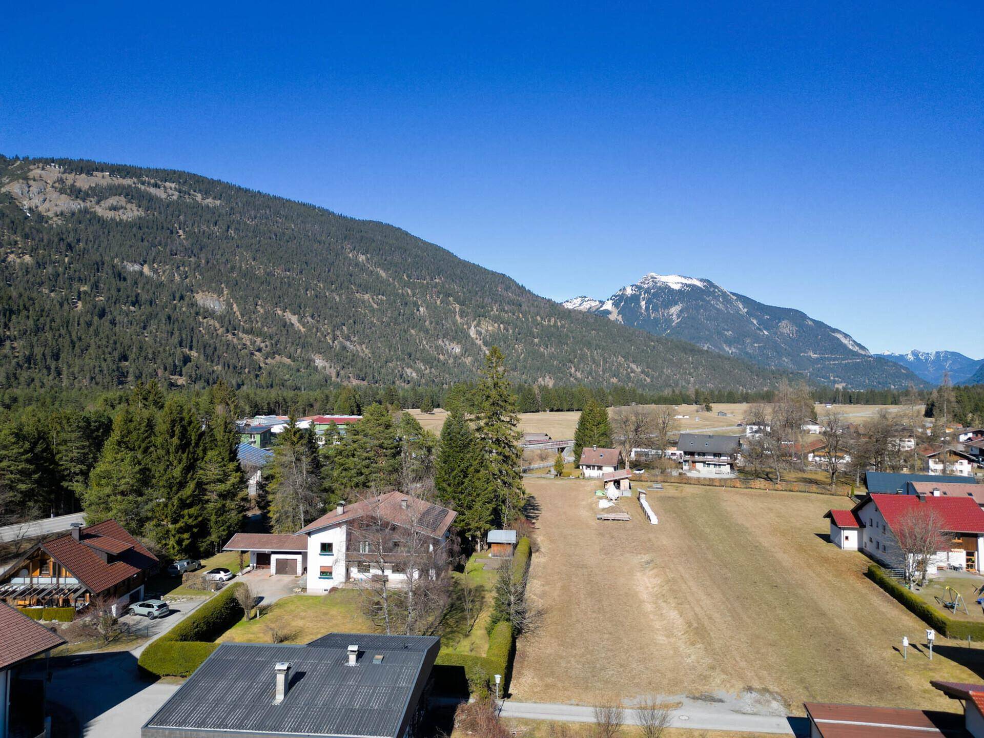 Luftaufnahme eines Dorfes mit Häusern, Feldern und majestätischen Bergen im Hintergrund unter blauem Himmel.