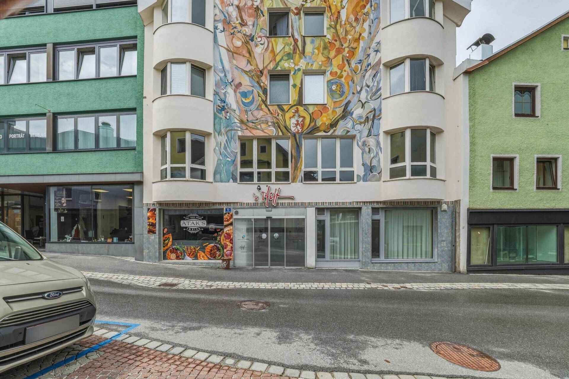 Eine belebte Straßenansicht mit einem Gebäude, das eine farbenfrohe Wandmalerei und Geschäfte im Erdgeschoss zeigt.