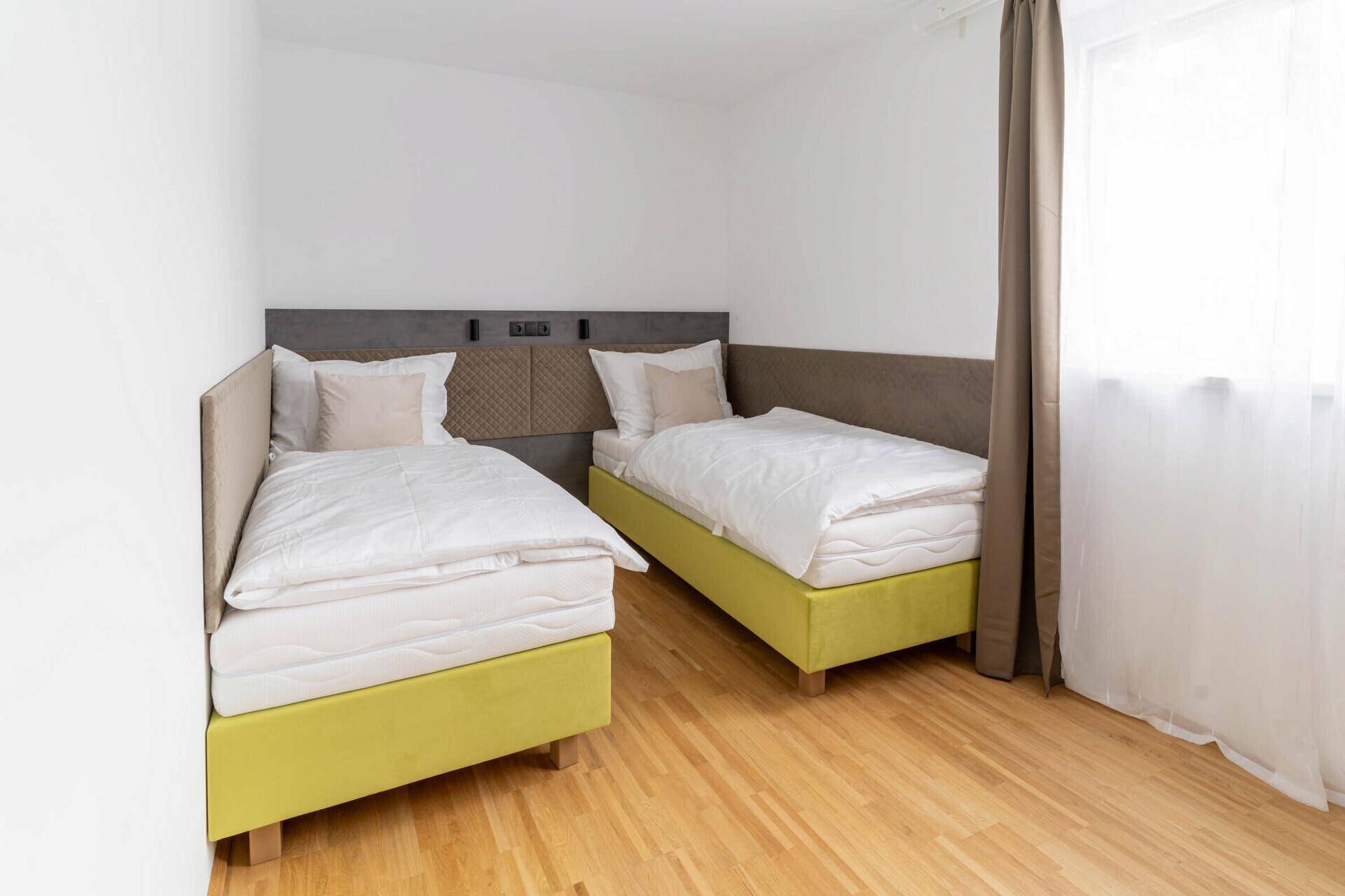 Helles Schlafzimmer mit zwei Einzelbetten, Holzboden und Fenster mit Vorhängen.