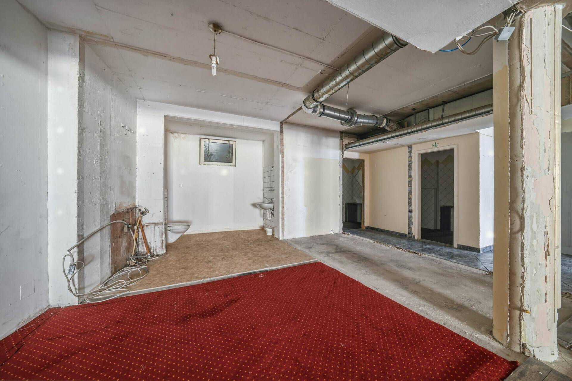 Leerstehender Gewerberaum mit rotem Teppich, sichtbaren Rohren und einem hinteren Bereich mit Toilette.