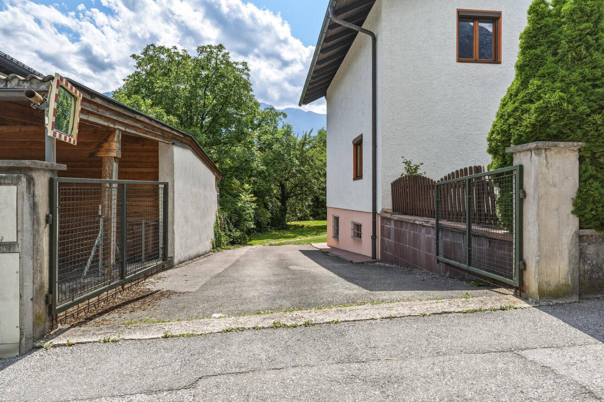 Zufahrt zur Garage neben dem Haus, umgeben von einem Zaun und Bäumen.