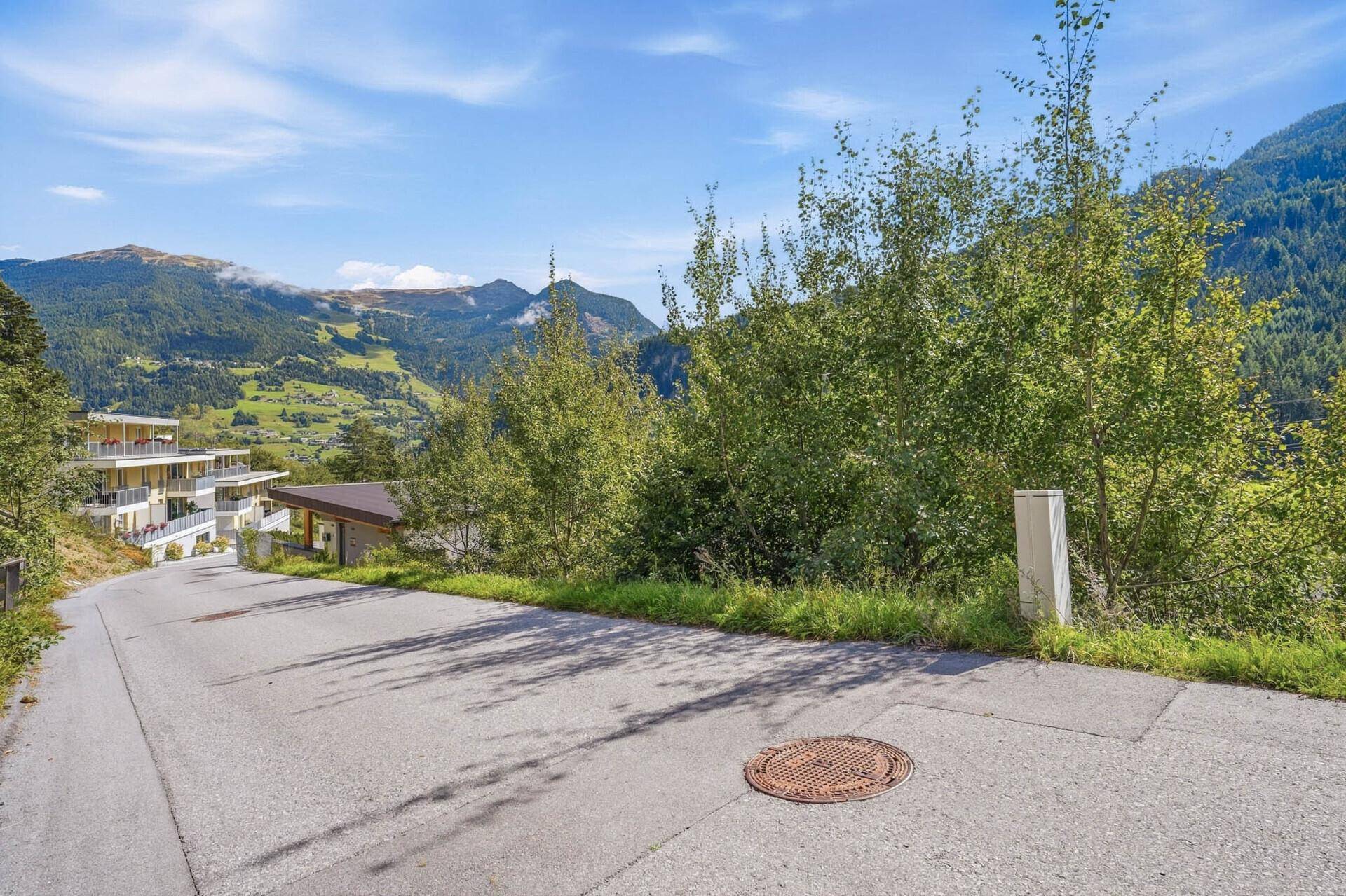 Landstraße mit grünem Randstreifen und Bäumen, im Hintergrund sind Häuser und Berge sichtbar.