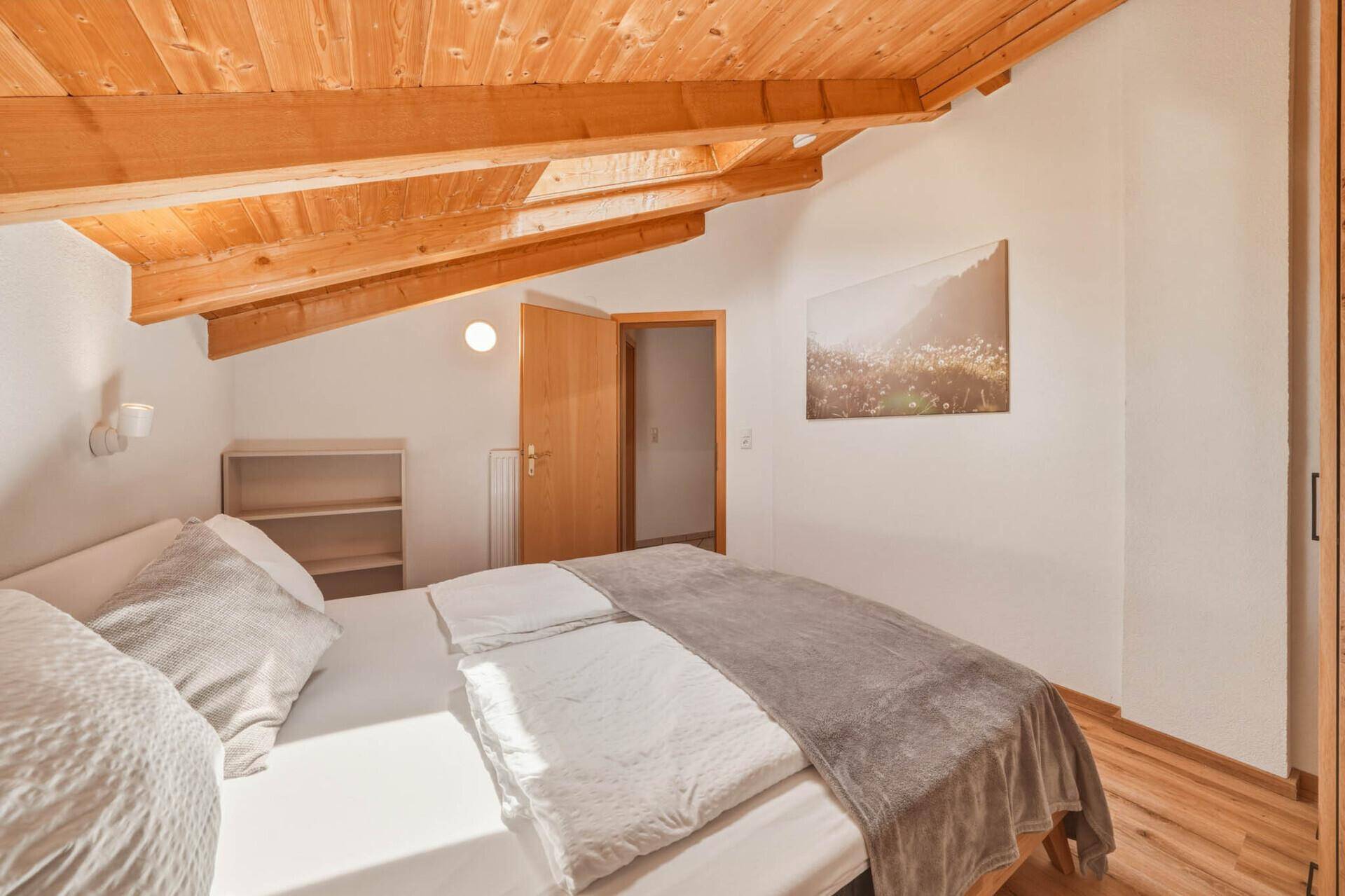 Helles Schlafzimmer mit Holzbalkendecke, Holzboden und einem Bild von Berglandschaft an der Wand.