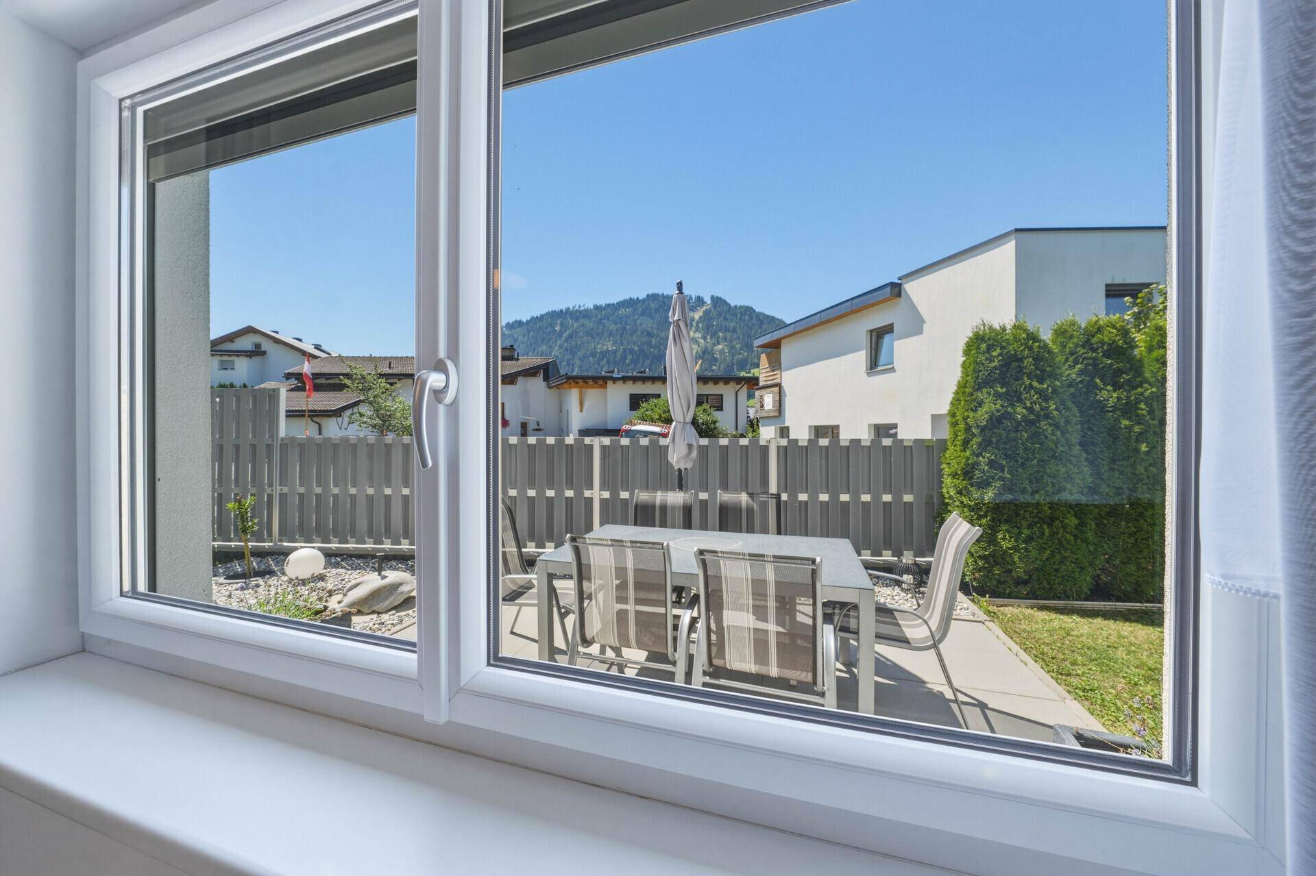 Blick aus dem Fenster auf die sonnige Terrasse mit Gartenmöbeln und Bergblick.