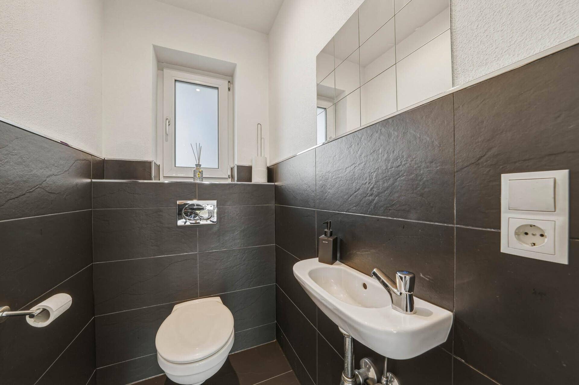 Modernes Gäste-WC mit dunklen Fliesen, wandhängendem WC, kleinem Waschbecken und Fenster.