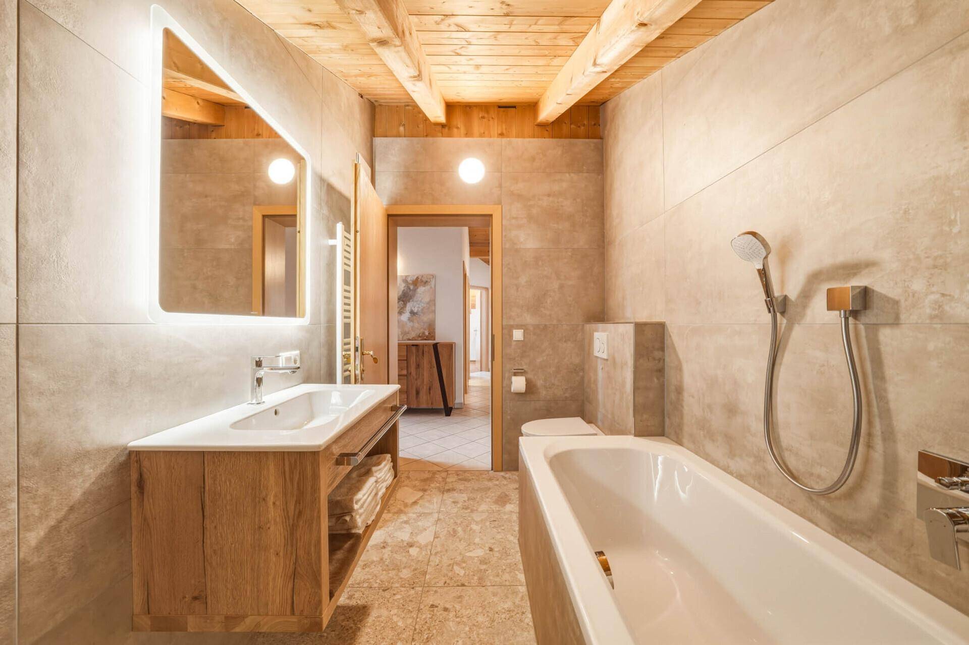 Geräumiges Badezimmer mit Badewanne, Holzbalkendecke, modernen Fliesen und beleuchtetem Spiegel.