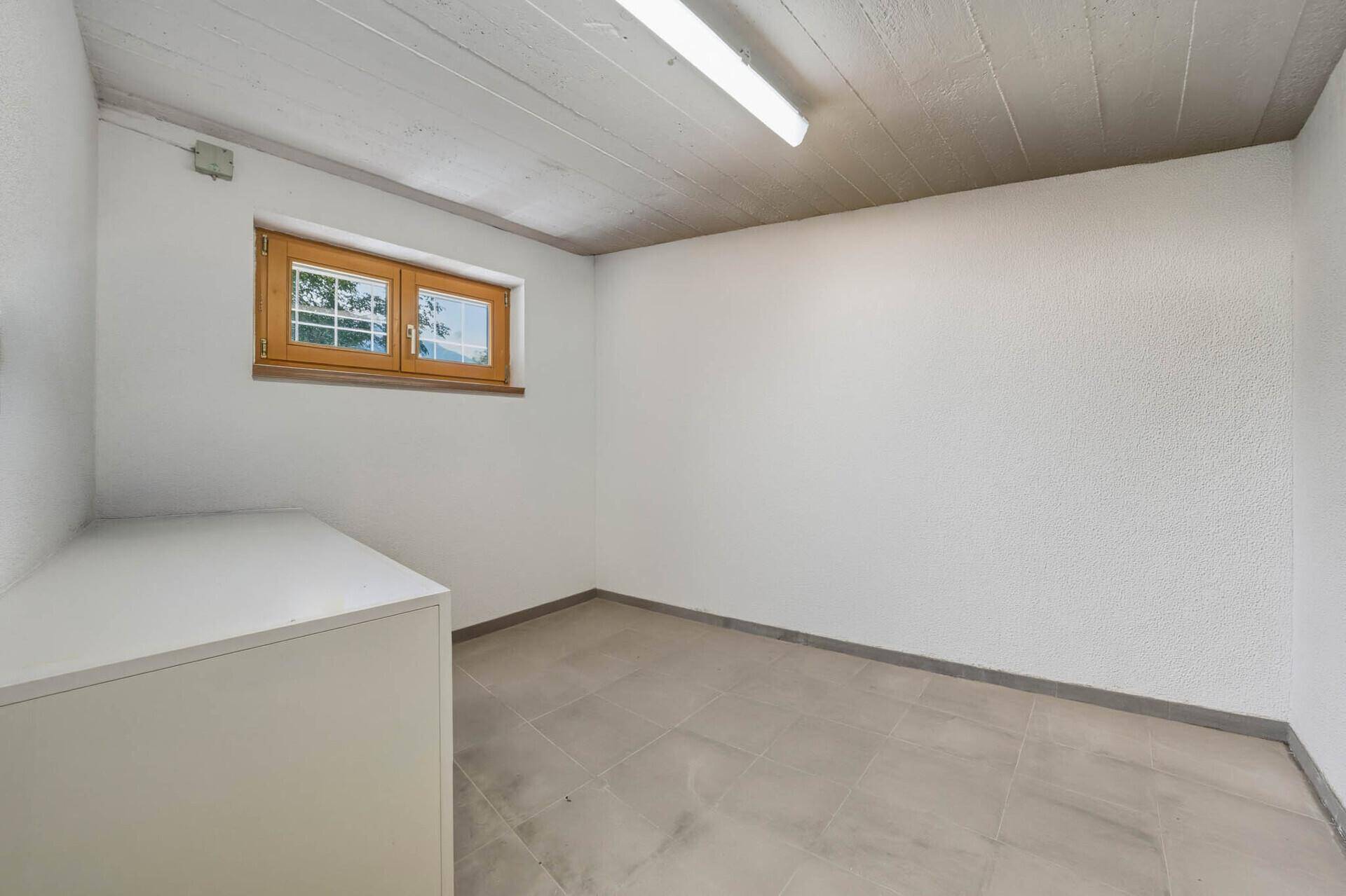 Leerer Kellerraum mit weißen Wänden, grauen Fliesen und einem kleinen Fenster.