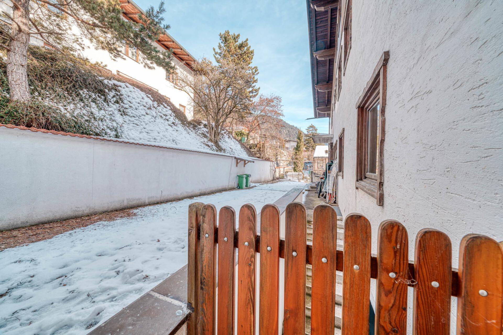 Seitlicher Weg mit Holzzaun und verschneiter Böschung neben dem Gebäude.