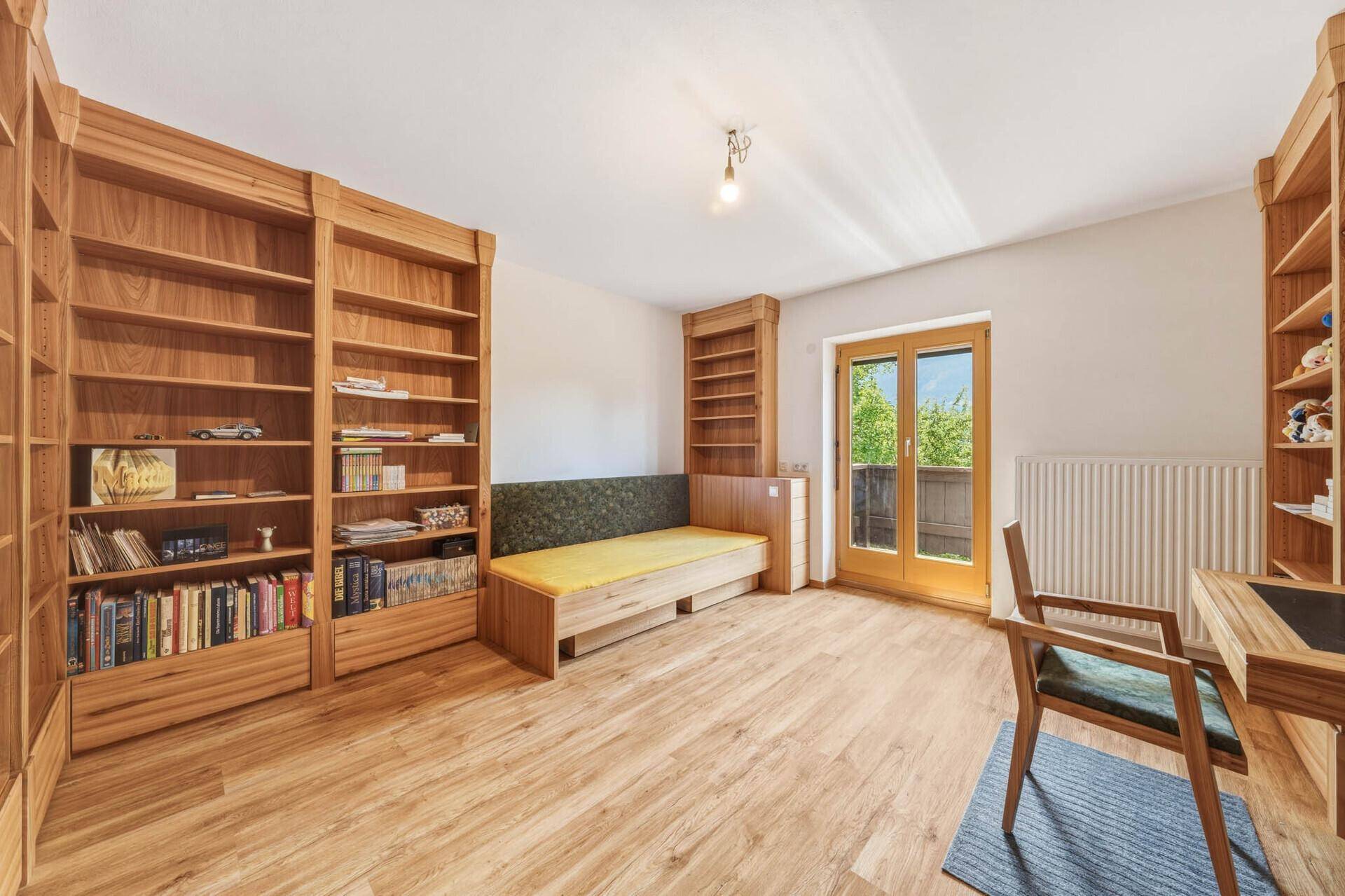 Helles Arbeitszimmer mit Holzbücherregalen, Schreibtisch und Zugang zu einem Außenbereich.