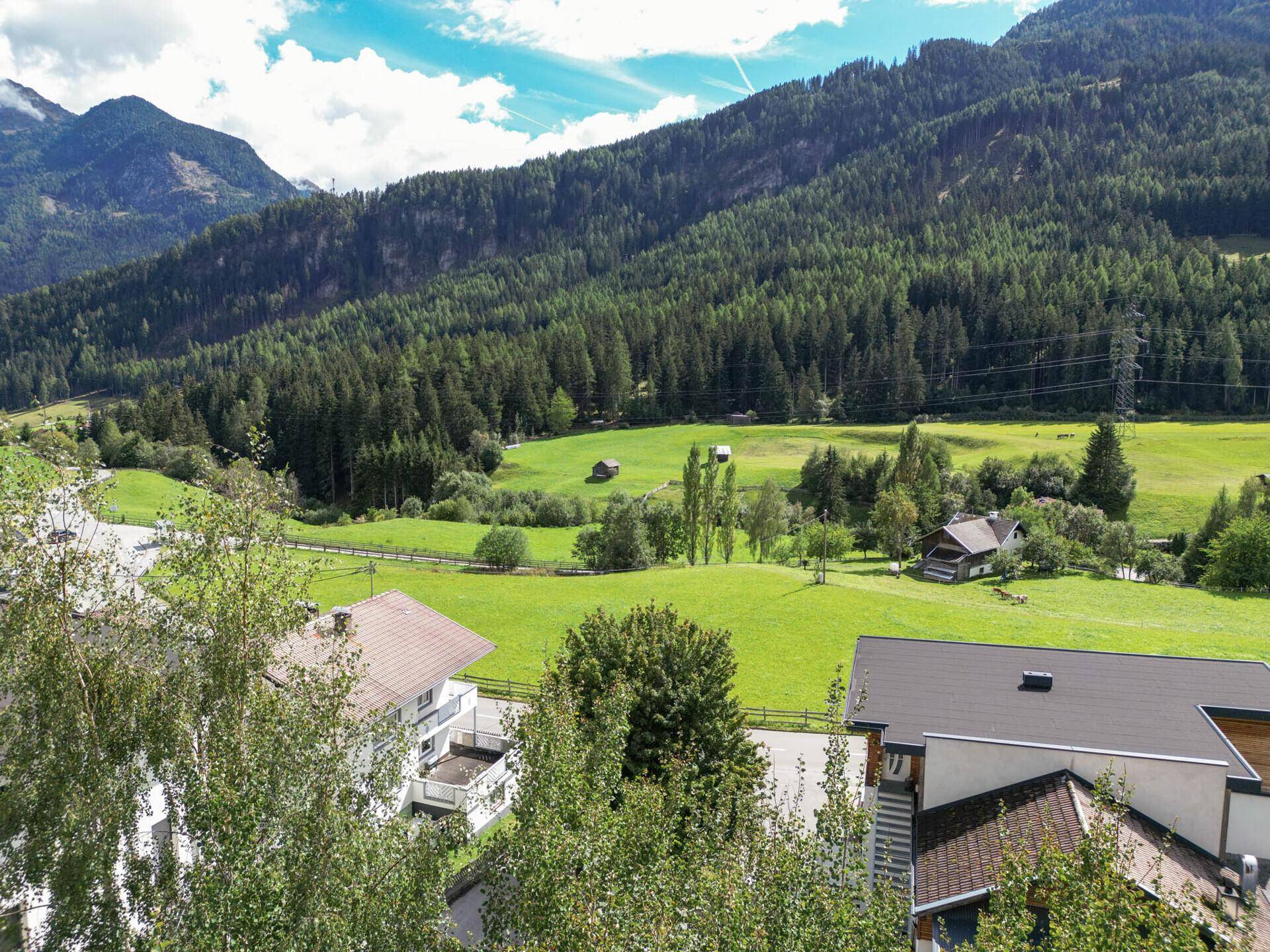 Panoramablick auf eine ländliche Landschaft mit Häusern, weiten Wiesen und dichten Wäldern an Berghängen.