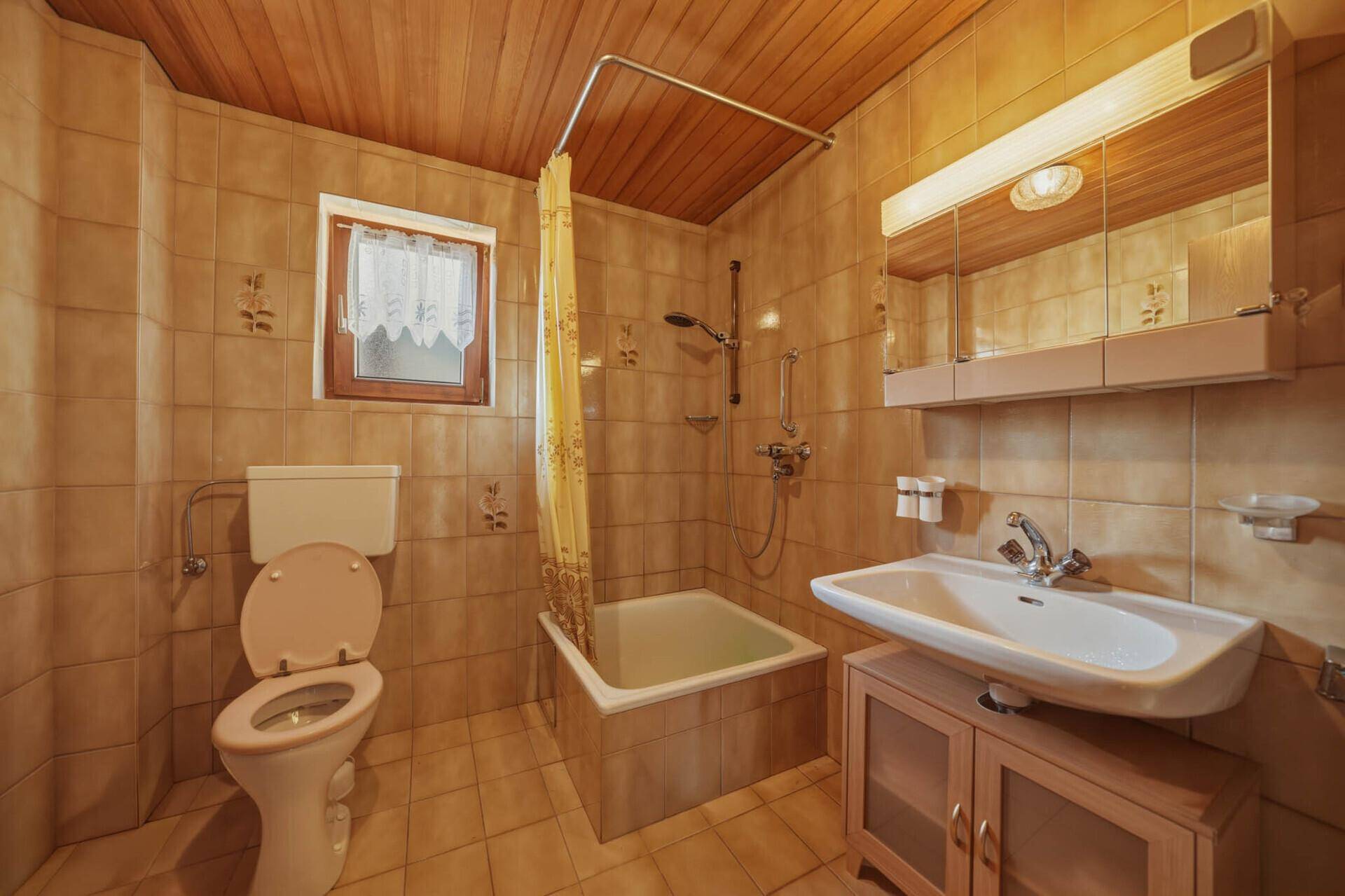 Badezimmer mit Badewanne, WC und Waschbecken, ausgestattet mit braunen Fliesen und Holzdecke.