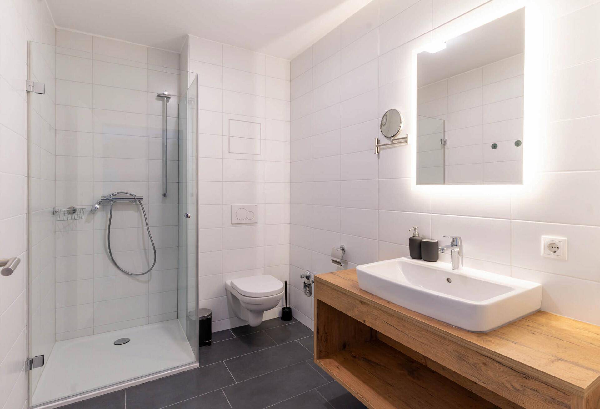 Modernes Badezimmer mit begehbarer Dusche, Holzwaschtisch und beleuchtetem Spiegel.