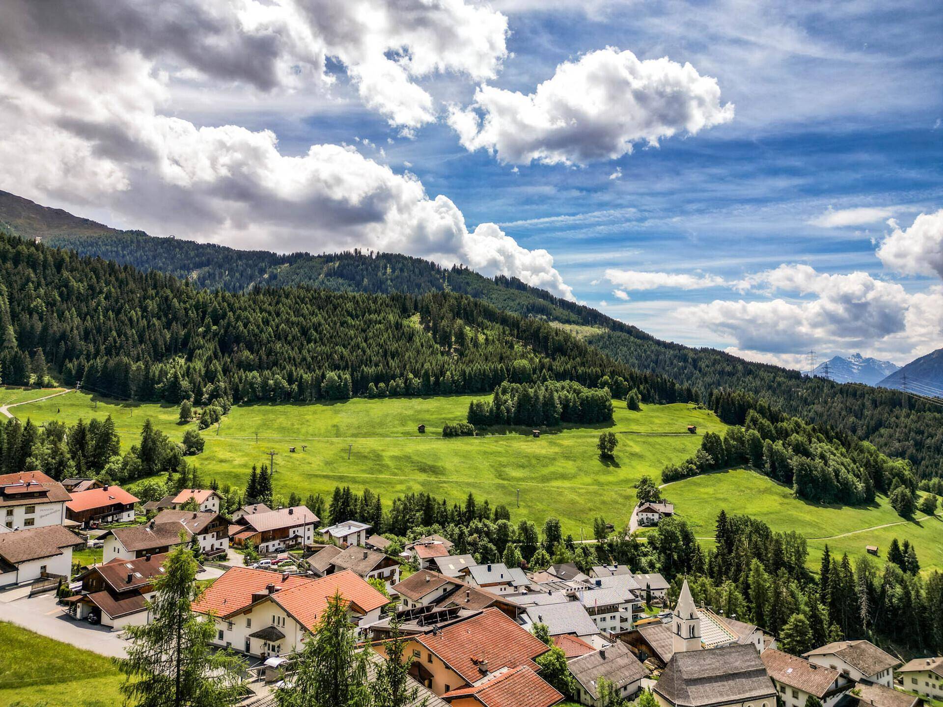 Weitläufiger Blick über ein idyllisches Dorf mit Kirche, eingebettet in eine grüne Berglandschaft.