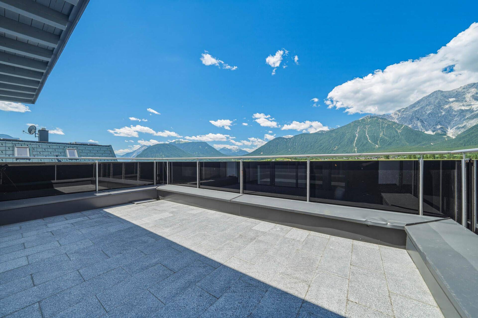 Große Dachterrasse mit Steinplatten und Glasgeländer, bietet einen weiten Bergblick.