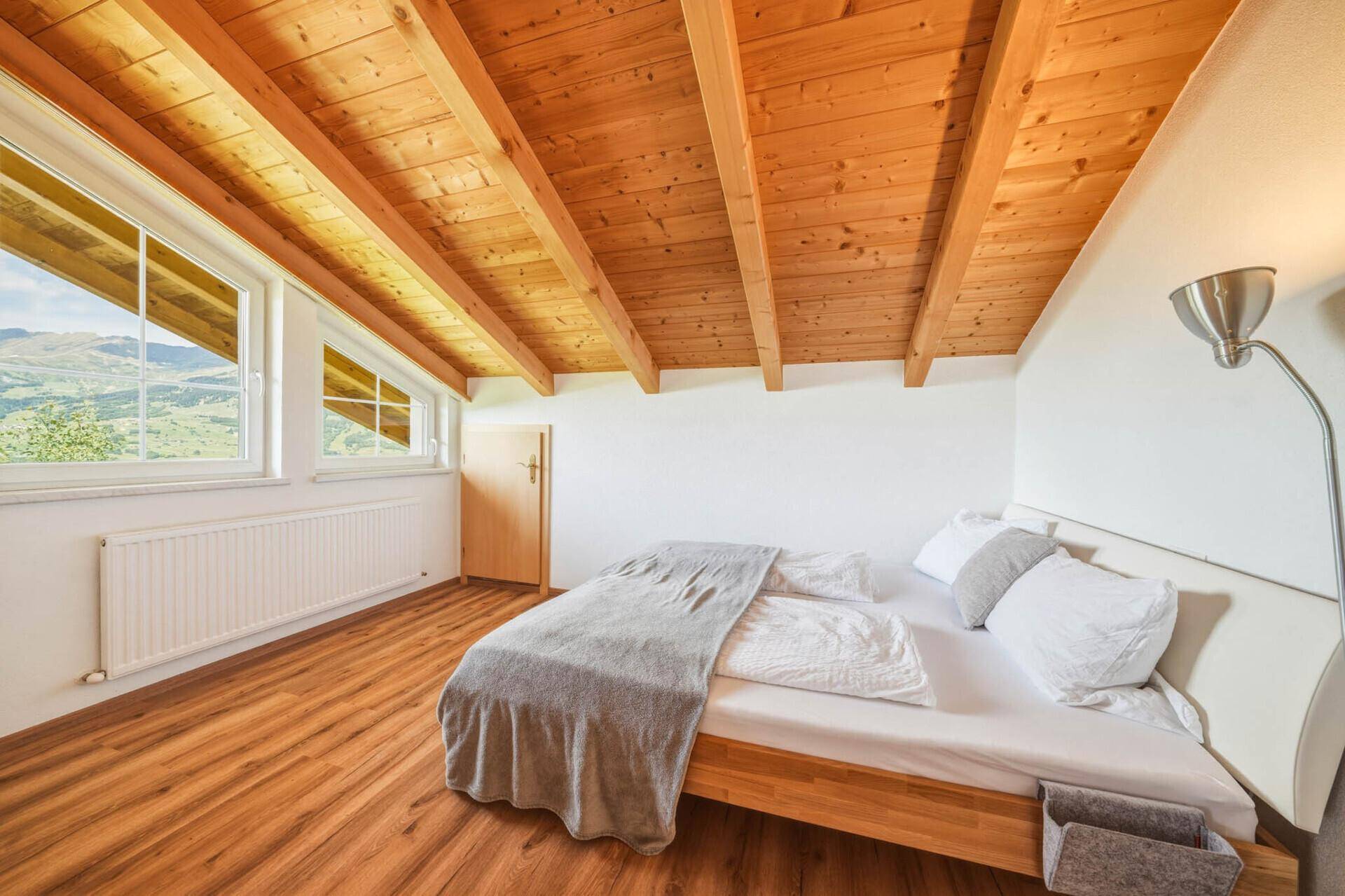 Geräumiges Schlafzimmer im Dachgeschoss mit Holzbalkendecke, Holzboden und großen Fenstern mit Bergblick.