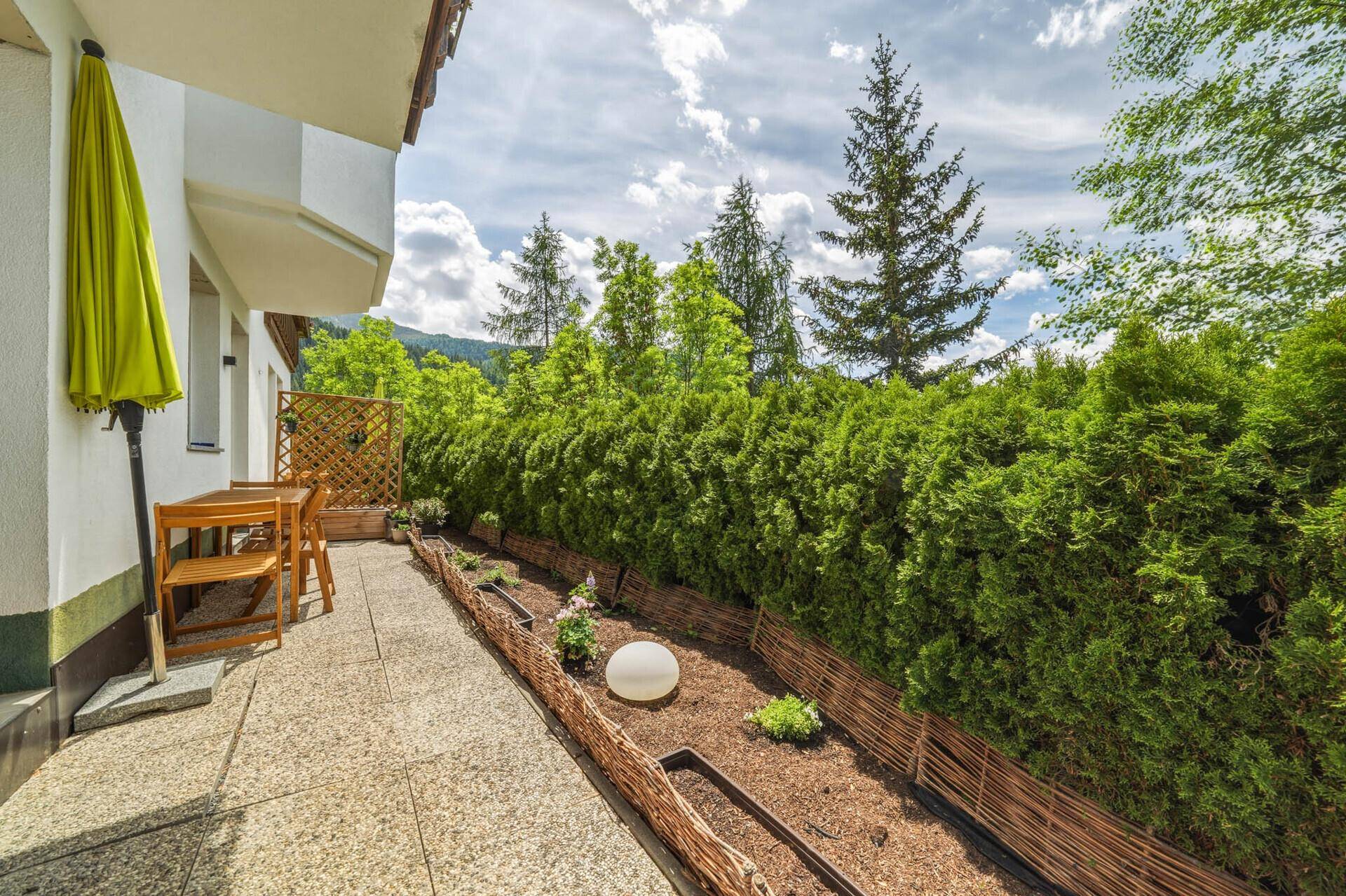 Lange Terrasse mit Gartenmöbeln und Sonnenschirm, umgeben von üppiger grüner Hecke und Pflanzen.