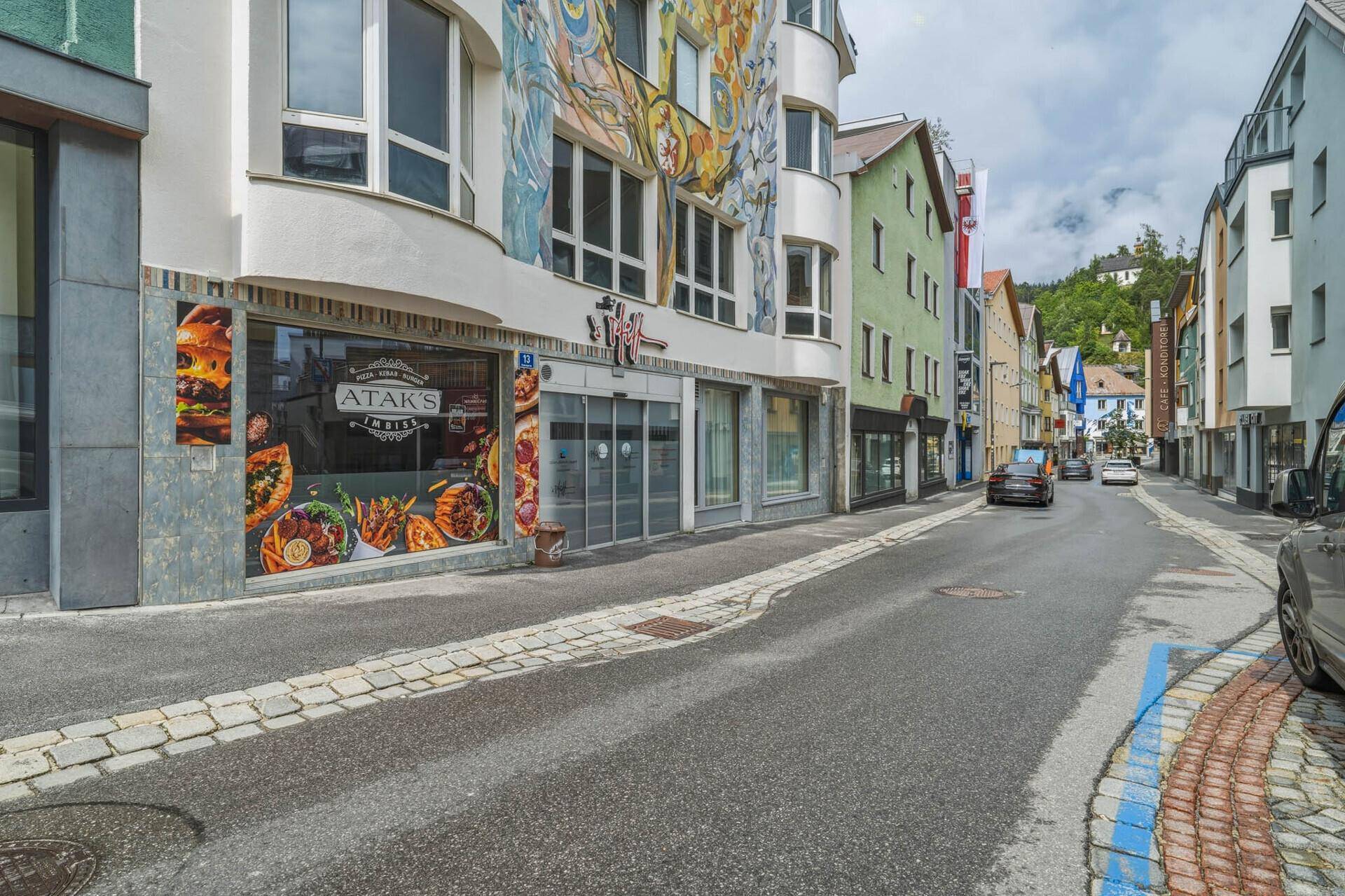 Eine Straße mit Geschäften im Erdgeschoss und einem Gebäude mit einer auffälligen Wandmalerei unter bewölktem Himmel.