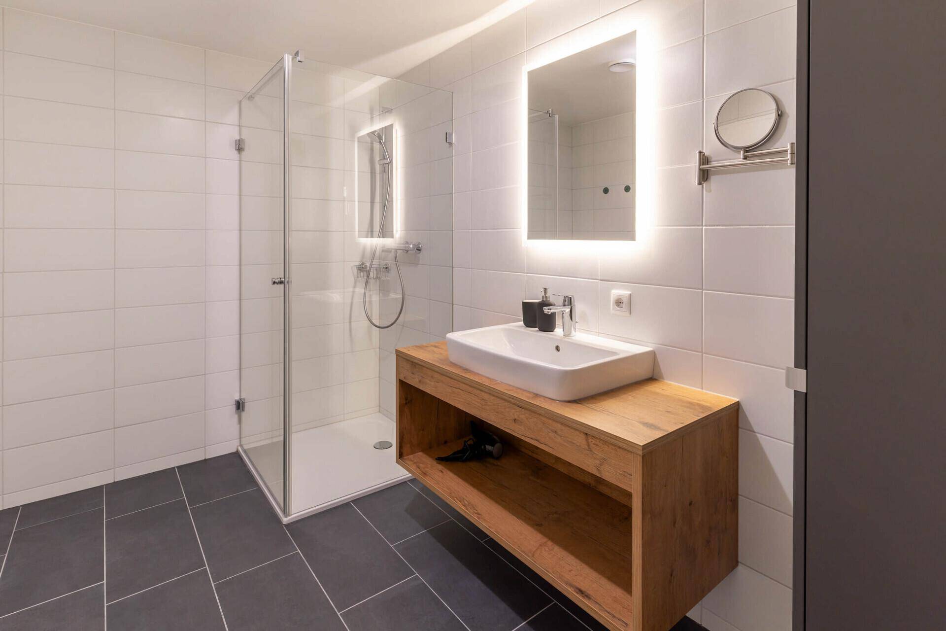 Modernes Badezimmer mit begehbarer Dusche, Holzwaschtisch und beleuchtetem Spiegel.