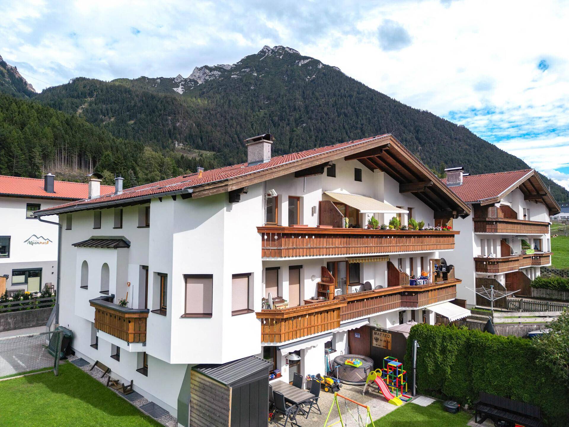 Alpines Mehrfamilienhaus mit Holzbalconen und einem kleinen Gartenbereich mit Spielplatz.