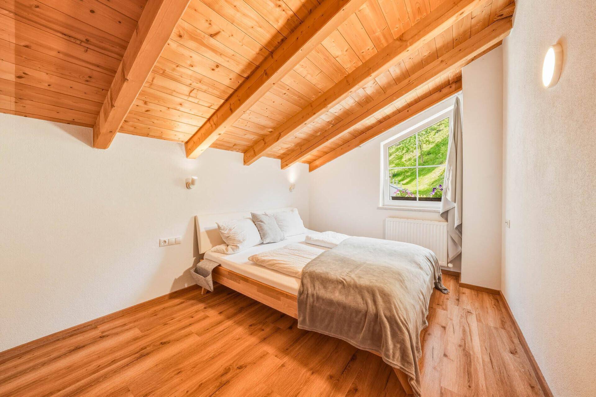 Schlafzimmer im Dachgeschoss mit Holzbalkendecke, Holzboden und Fenster mit Blick ins Grüne.