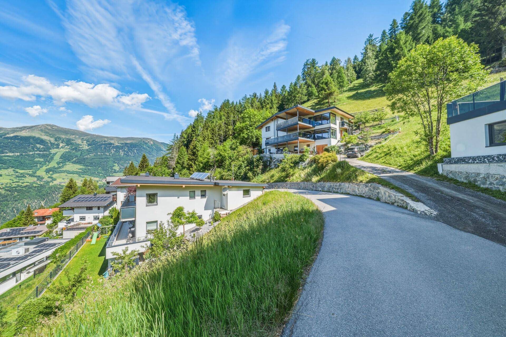 Panoramablick auf moderne Häuser entlang einer Bergstraße mit atemberaubender Alpenkulisse.