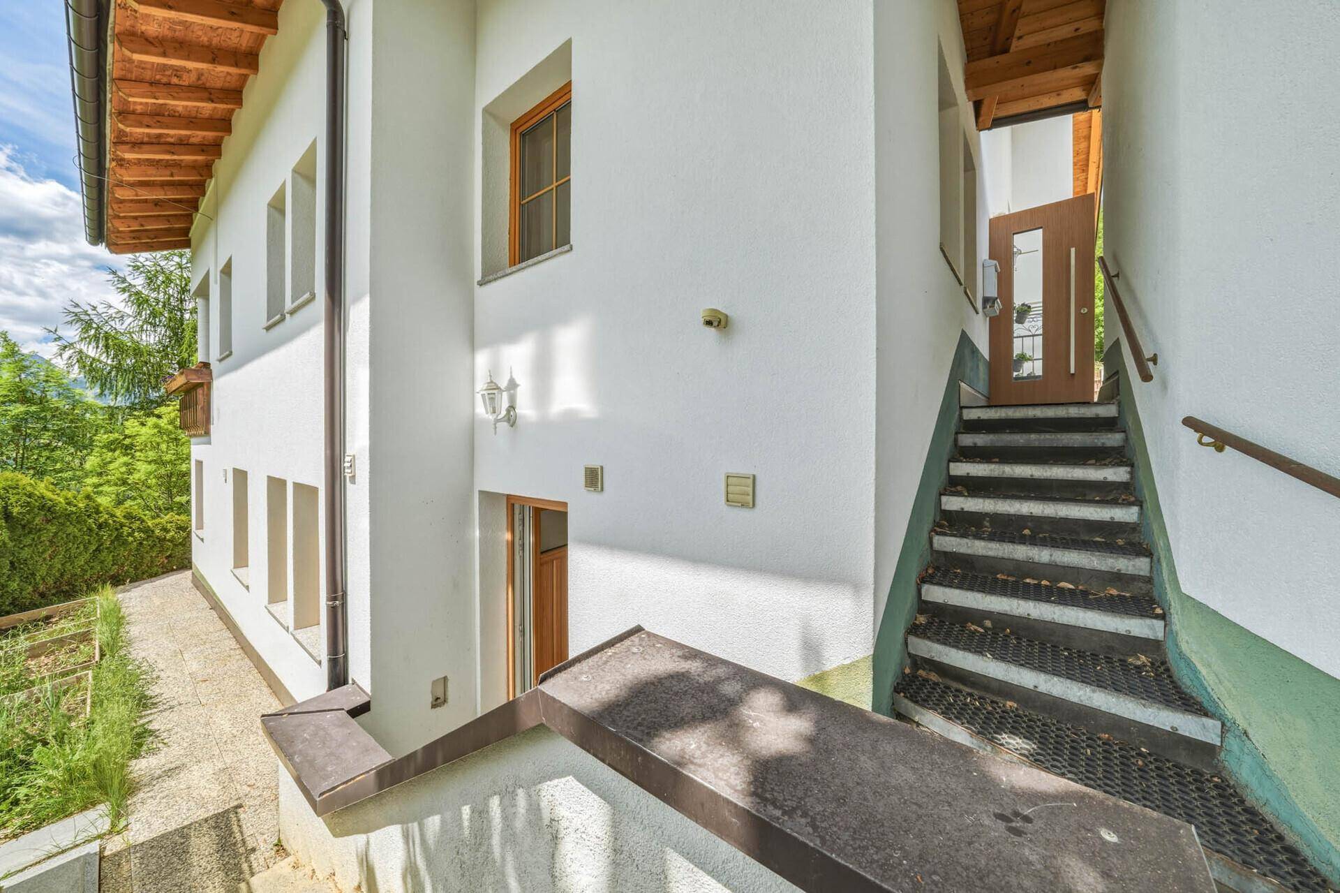 Außentreppe führt zum Eingang eines weißen Hauses mit Holzfenstern und grüner Umgebung.