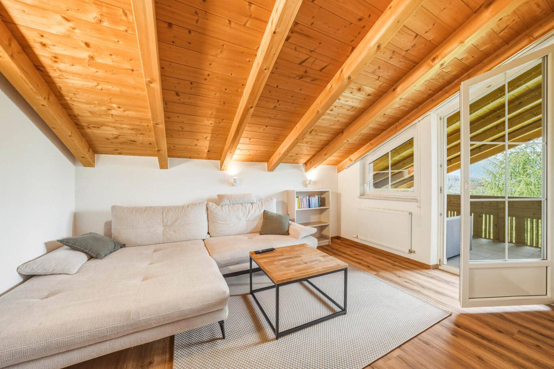 Gemütliches Wohnzimmer im Dachgeschoss mit Ecksofa, Holzbalkendecke und Zugang zum Balkon.