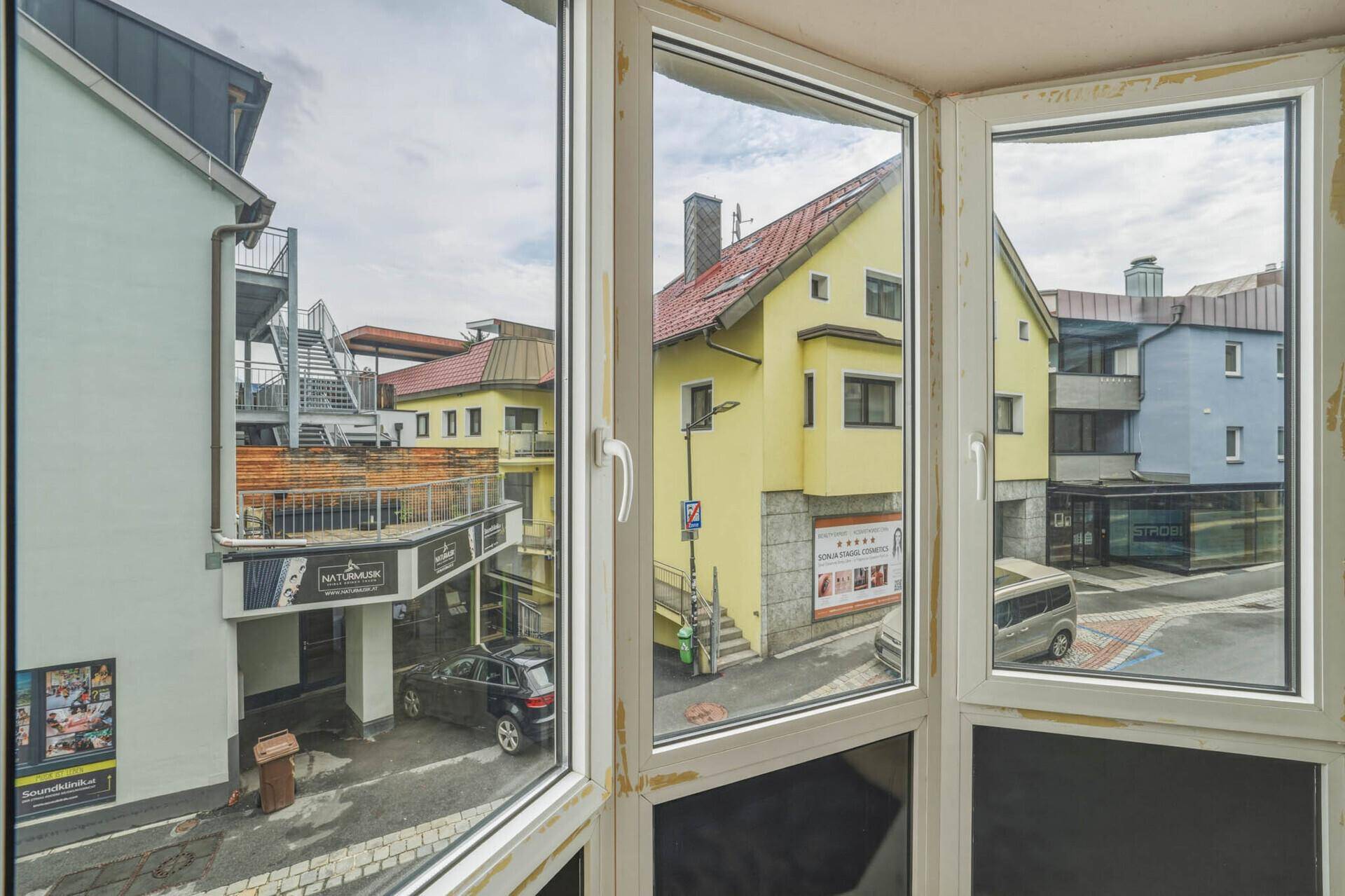 Blick aus einem Fenster auf eine Gasse mit einem gelben Gebäude und weiteren Häusern im Hintergrund.
