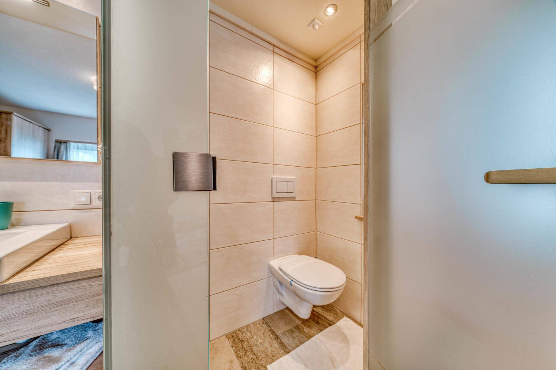 Modernes WC mit Wandtoilette und beigefarbenen Fliesen, angrenzend an einen Waschbereich.
