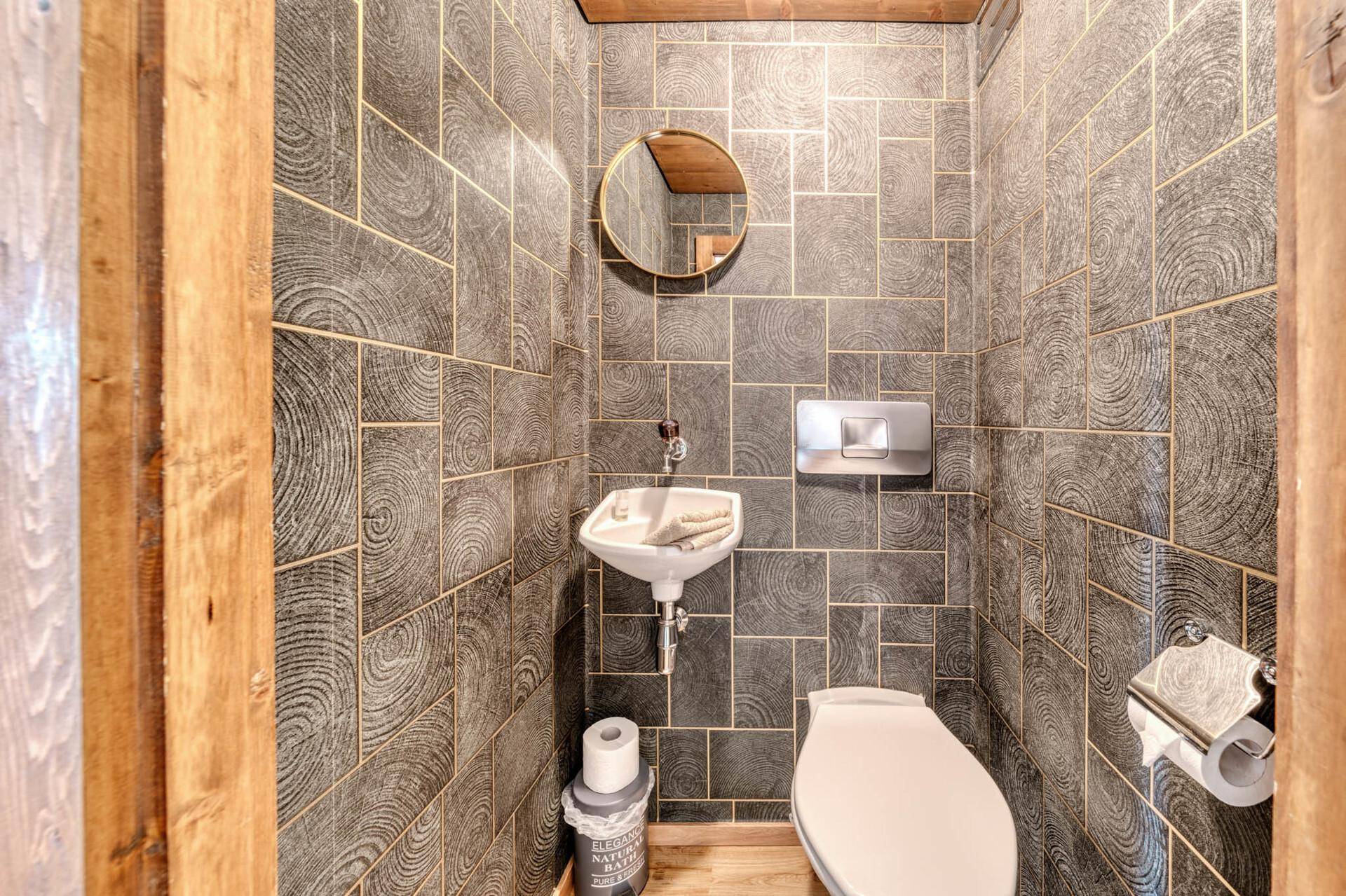 Modernes WC mit dunklen Fliesen in Holzoptik, Wandtoilette und kleinem Handwaschbecken.