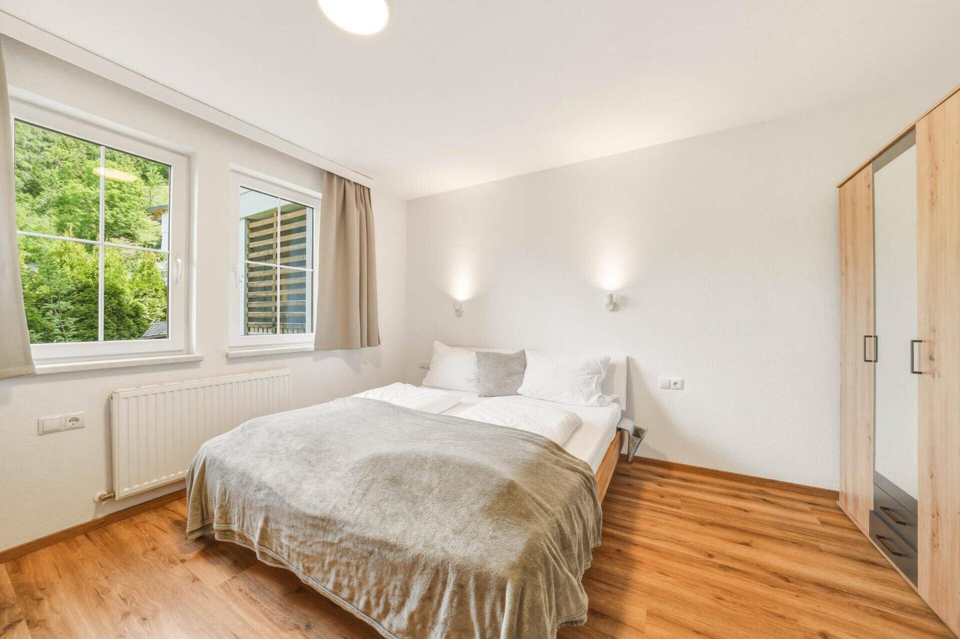 Helles Schlafzimmer mit Holzboden, Doppelbett und Fenstern mit Blick auf grüne Bäume.