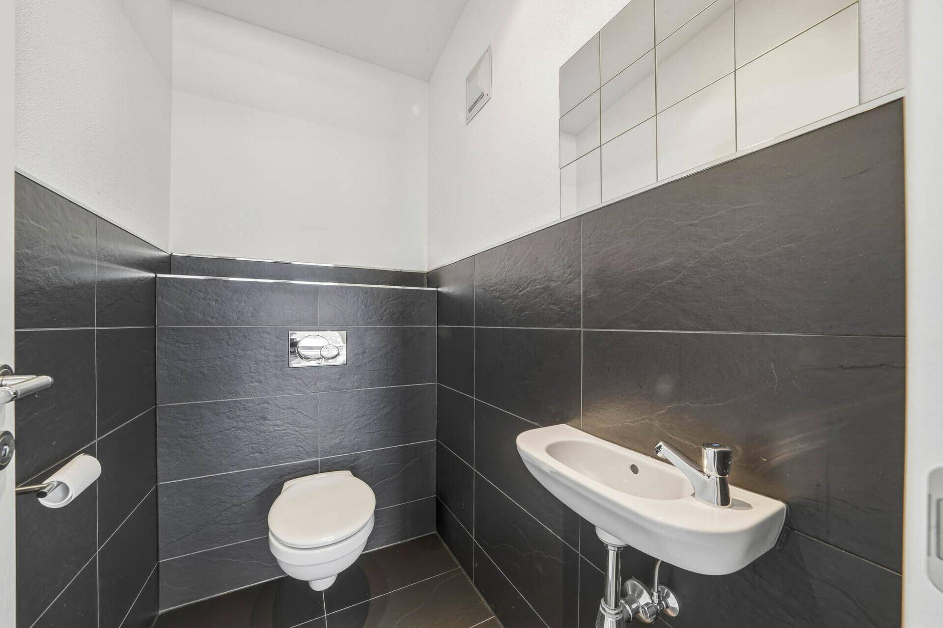 Modernes Gäste-WC mit Wand-WC, kleinem Waschbecken und dunklen Wandfliesen.