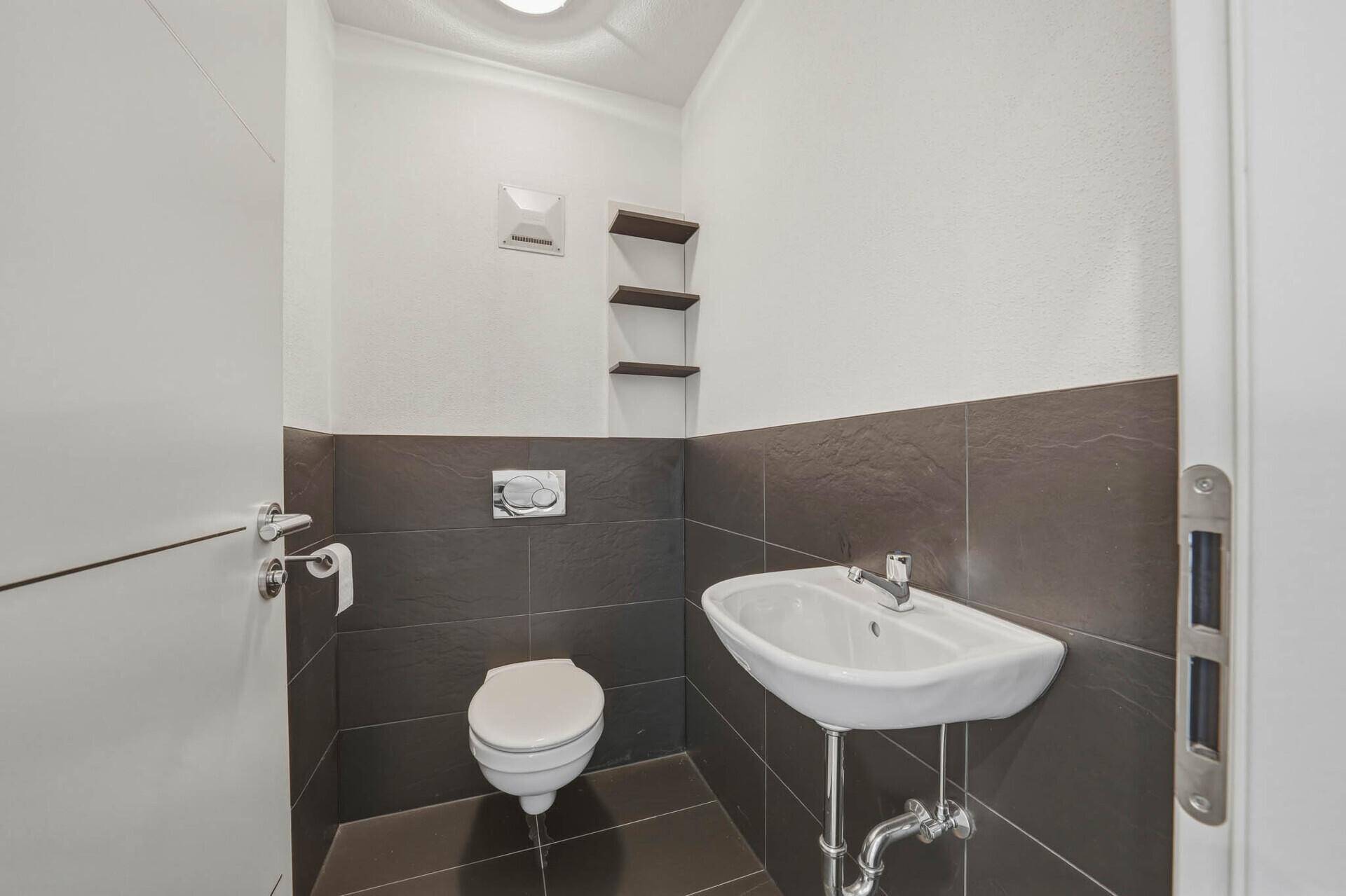 Kleines Gäste-WC mit Wand-WC, Waschbecken und dunklen Fliesen im unteren Wandbereich.