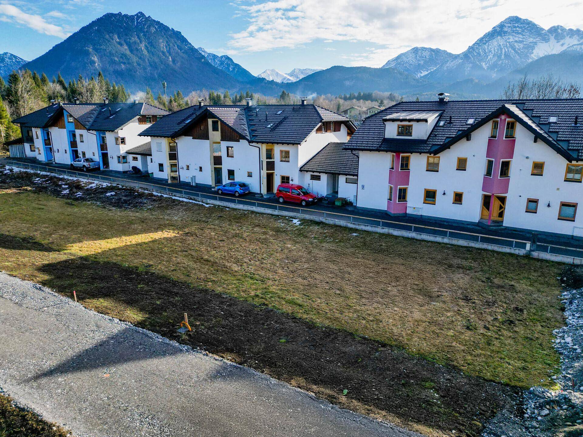 Reihenhäuser mit farbigen Fassaden und einem unbebauten Grundstück im Vordergrund, umgeben von Bergen.