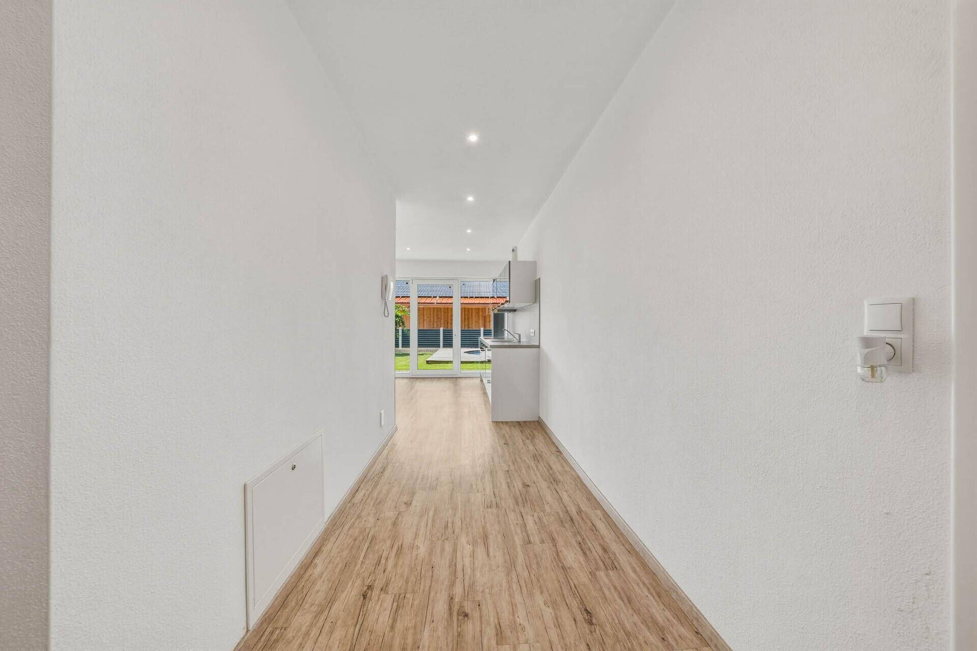 Langer Flur mit Laminatboden, der zum hellen, offenen Wohnbereich mit großen Fenstern führt.