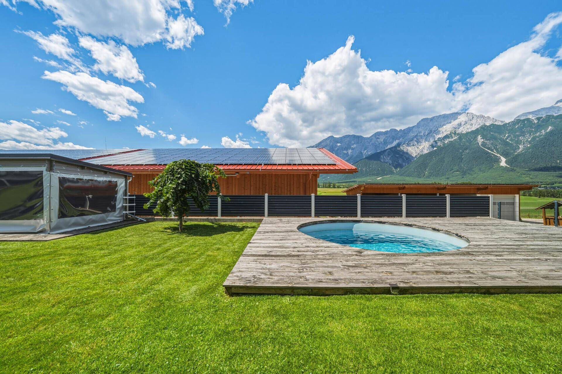 Gartenbereich mit Pool, Holzdeck und einem Gebäude mit Photovoltaikanlage vor Bergkulisse.