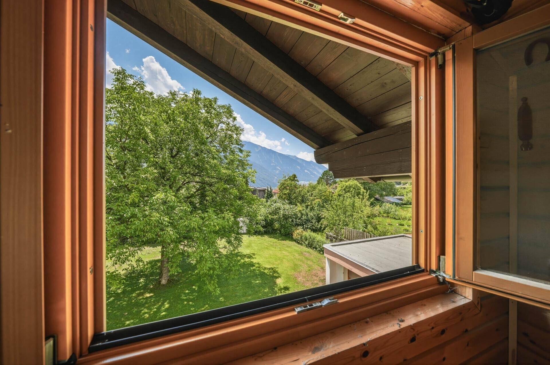 Blick aus einem Holzfenster auf einen grünen Garten und die umliegende Berglandschaft.