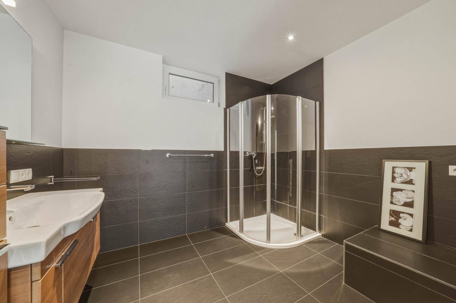 Badezimmer mit moderner Duschkabine, Waschtisch mit Holzunterschrank und dunklen Wandfliesen.