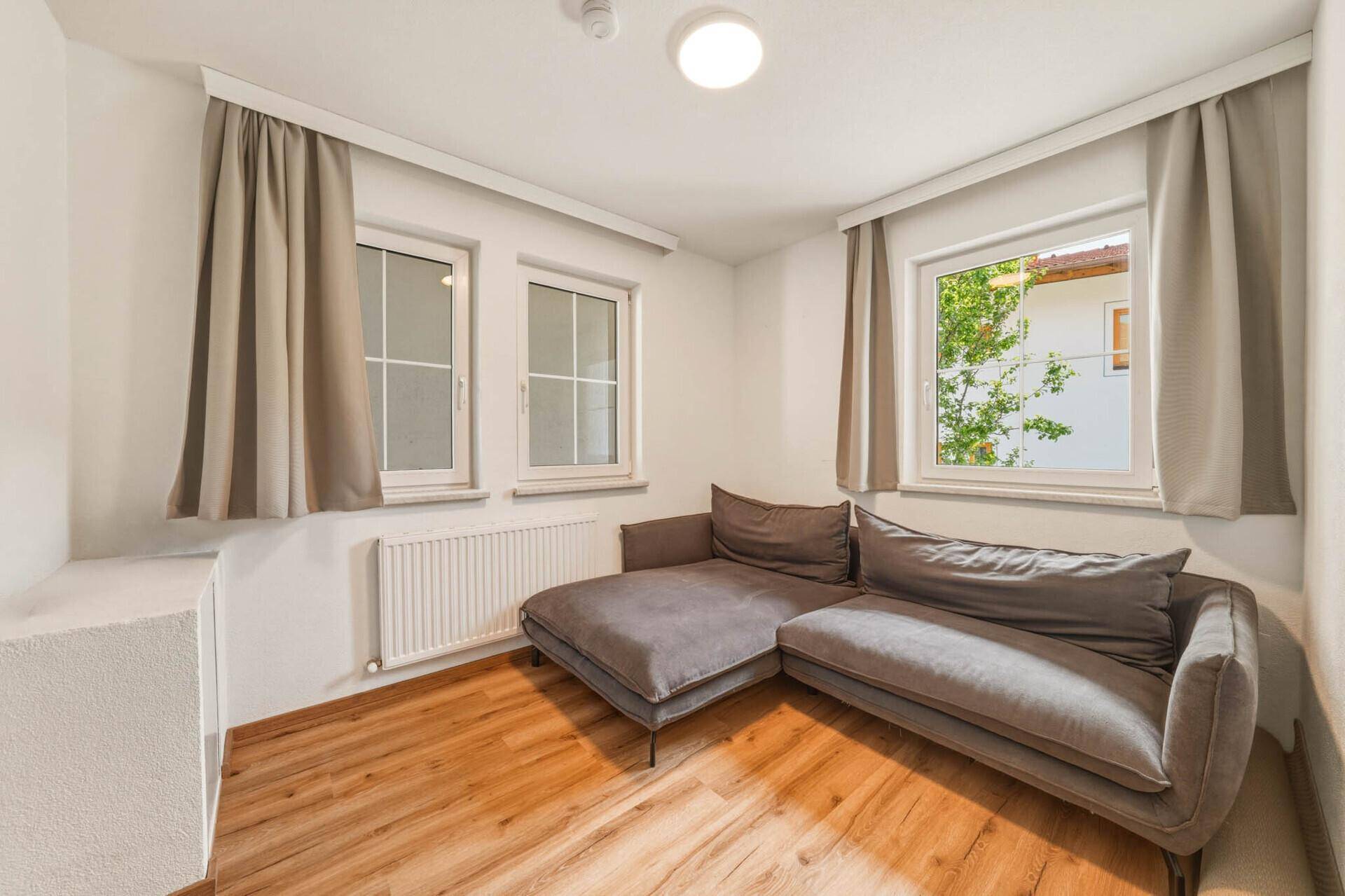 Gemütliches Zimmer mit grauem Ecksofa, Holzboden und hellen Fenstern.