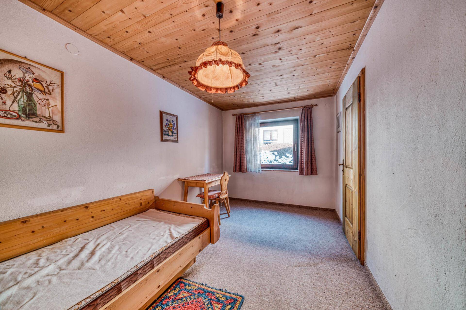 Einzelzimmer mit Holzdecke, Teppichboden und kleinem Schreibtisch am Fenster.