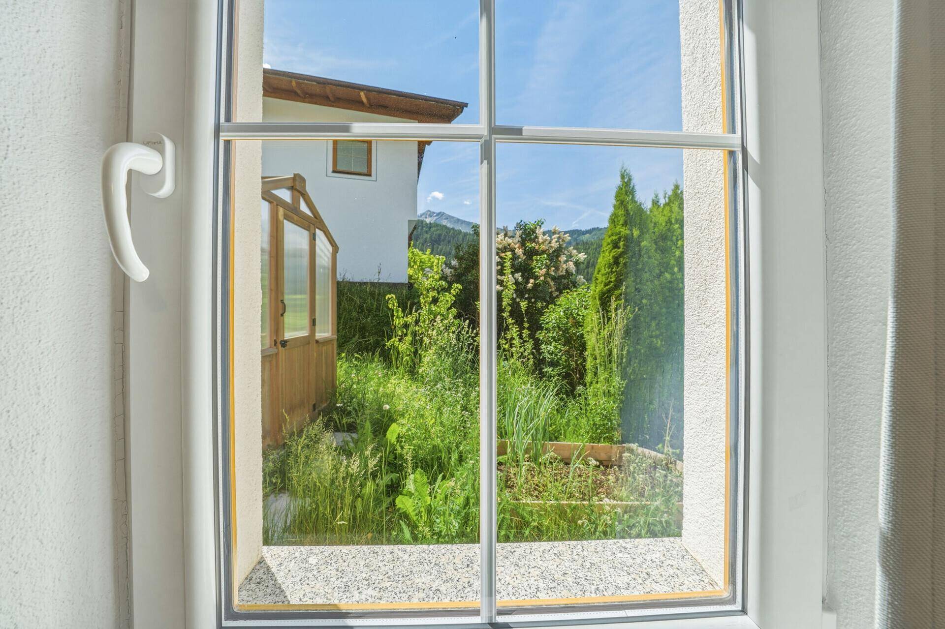 Fensterblick auf einen grünen Garten mit kleinem Gewächshaus und Bergsilhouette im Hintergrund.