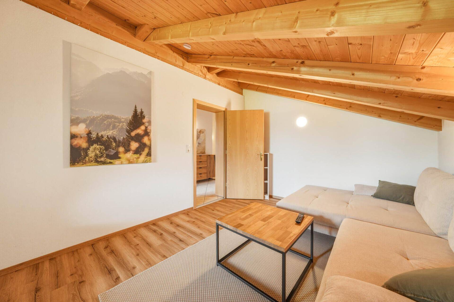 Wohnzimmer im Dachgeschoss mit Ecksofa, Holzbalkendecke und Blick auf ein Bergpanorama-Bild.