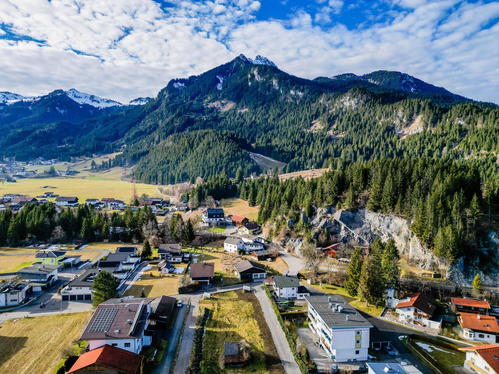 Panoramablick auf ein malerisches Dorf in den Bergen, umgeben von Wäldern und schneebedeckten Gipfeln.