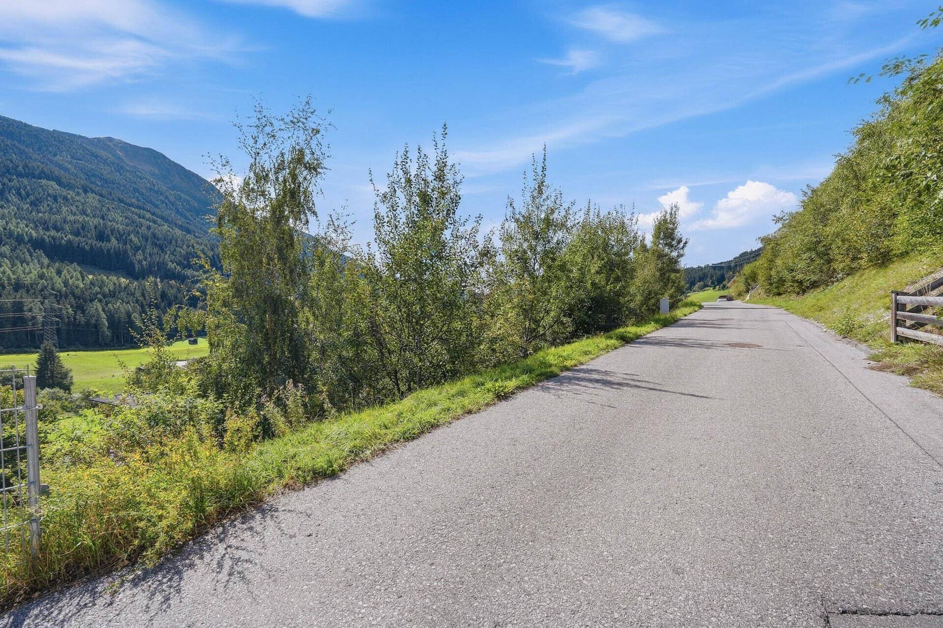Eine Landstraße mit Zaun und grünen Hängen, die sich durch ein Tal mit Bergen schlängelt.