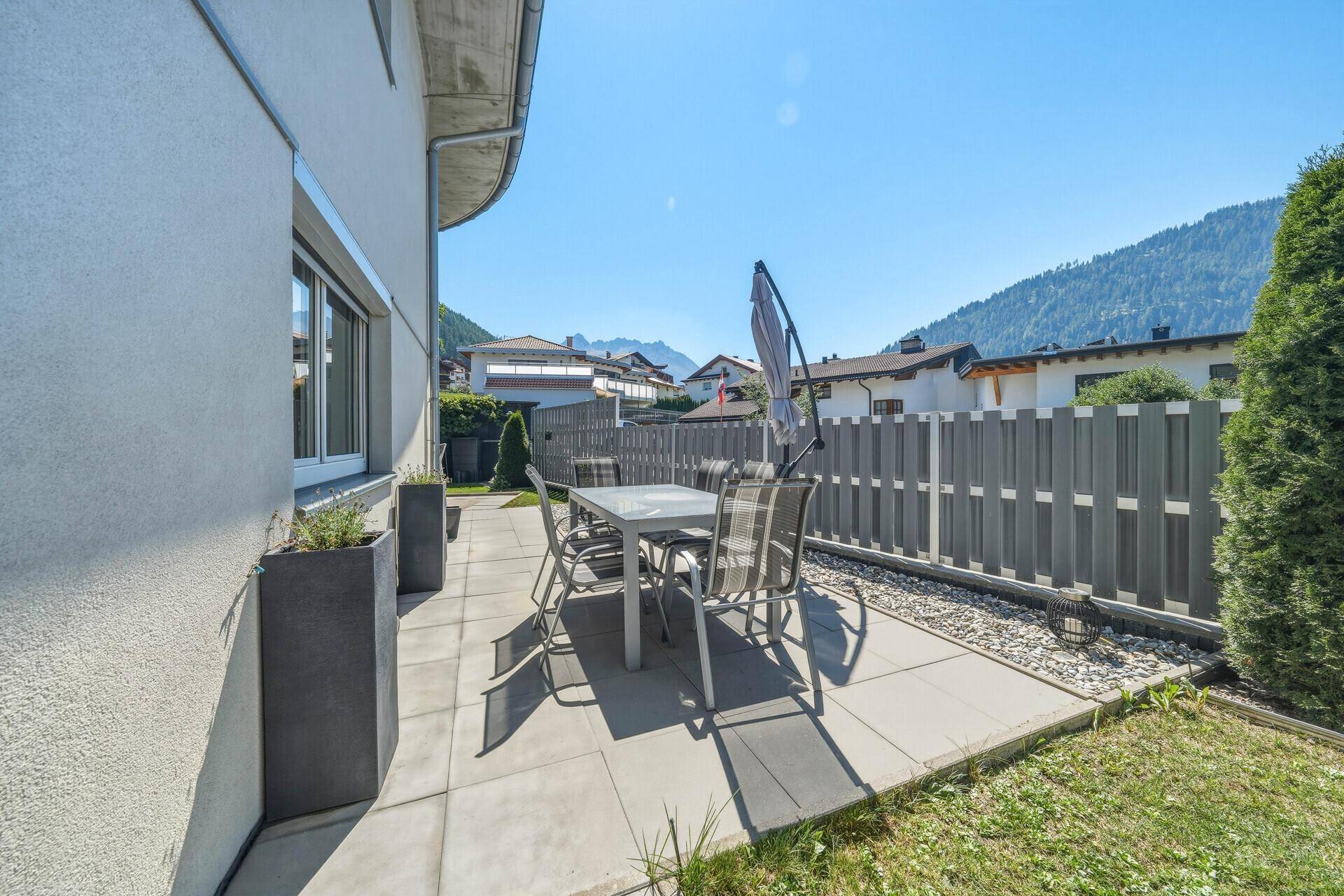 Helle Terrasse mit Esstisch, Sonnenschirm und weitem Blick auf die grünen Berge.