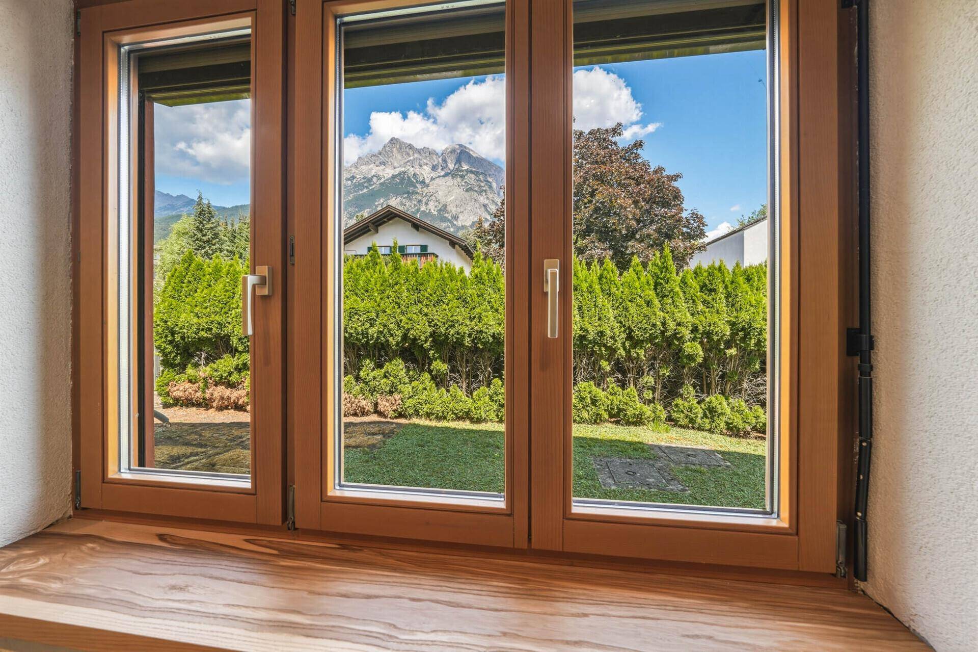 Fenster mit Blick auf den gepflegten Garten, eine Hecke und die umliegenden Berge.