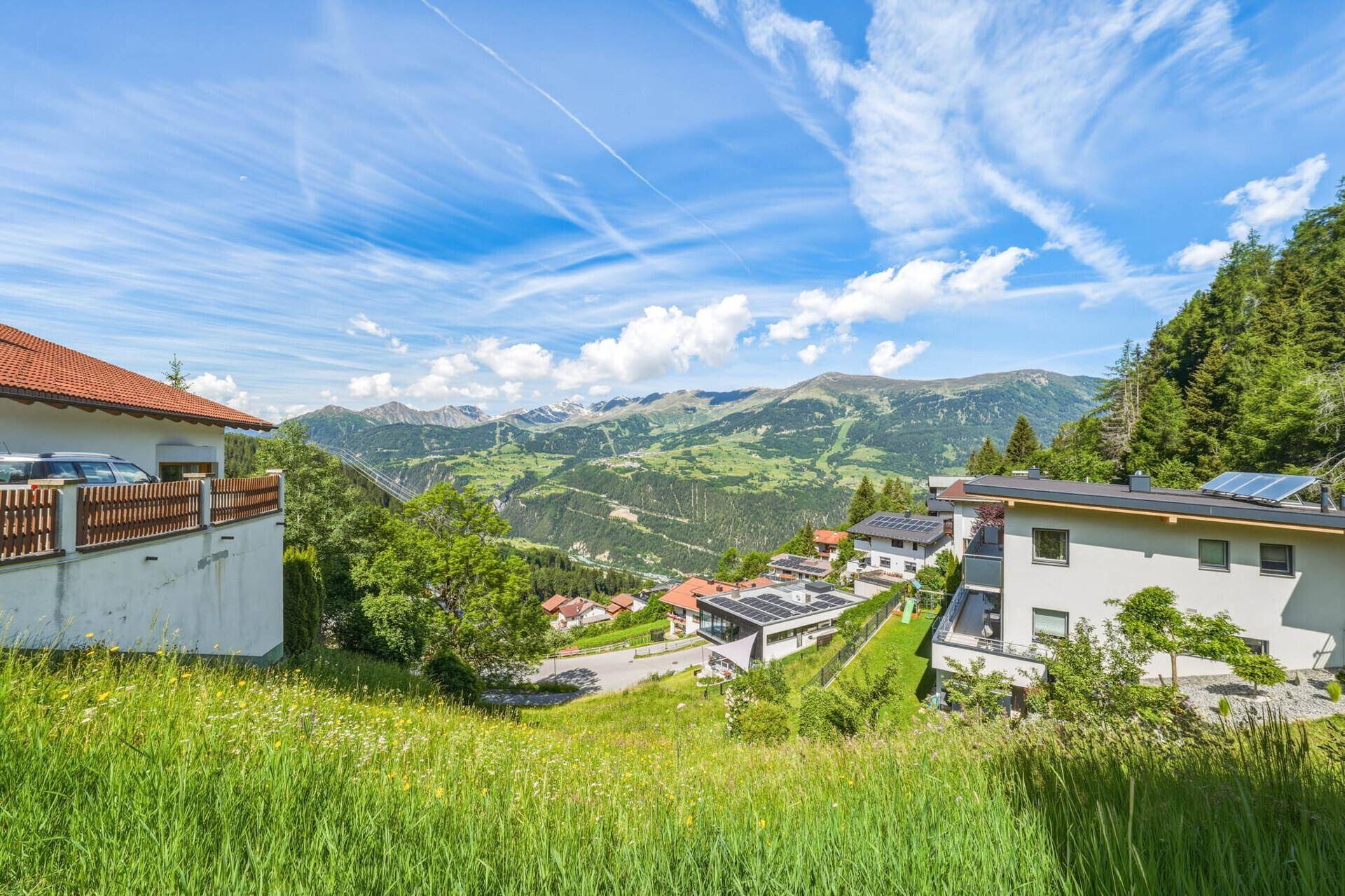 Weitläufiger Blick über grüne Hügel und eine Siedlung mit majestätischem Bergpanorama im Hintergrund.