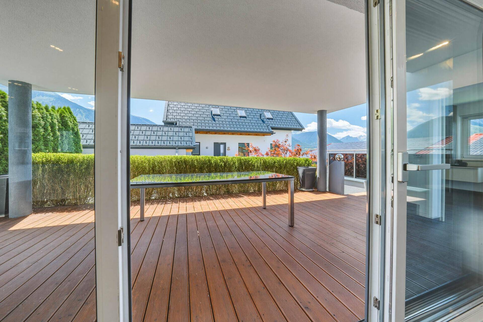 Große Holzterrasse mit Glas-Esstisch, umgeben von einer Hecke und mit Bergblick im Hintergrund.