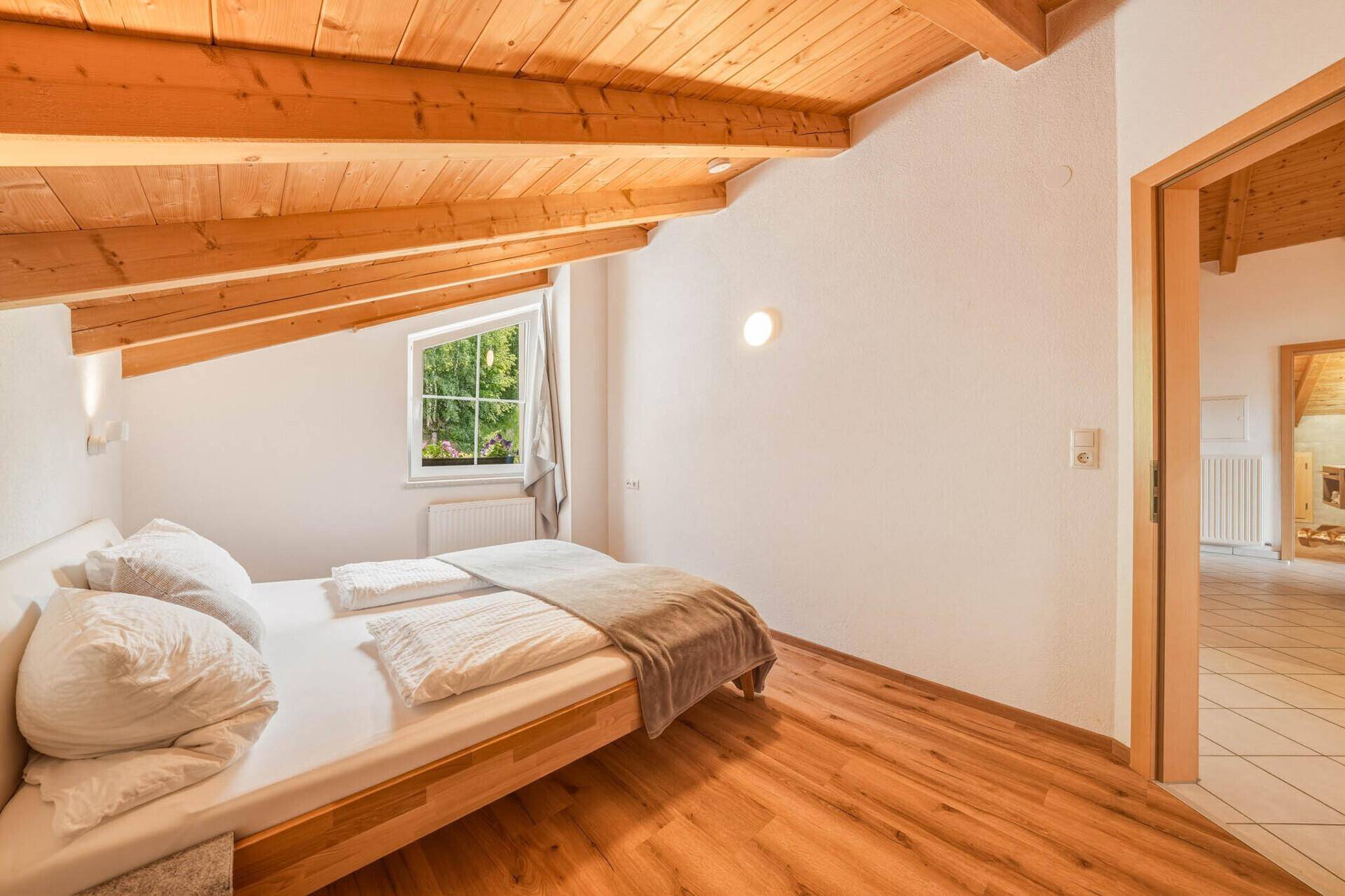 Helles Schlafzimmer im Dachgeschoss mit Holzbalkendecke, Holzboden und Fenster mit Blick ins Grüne.