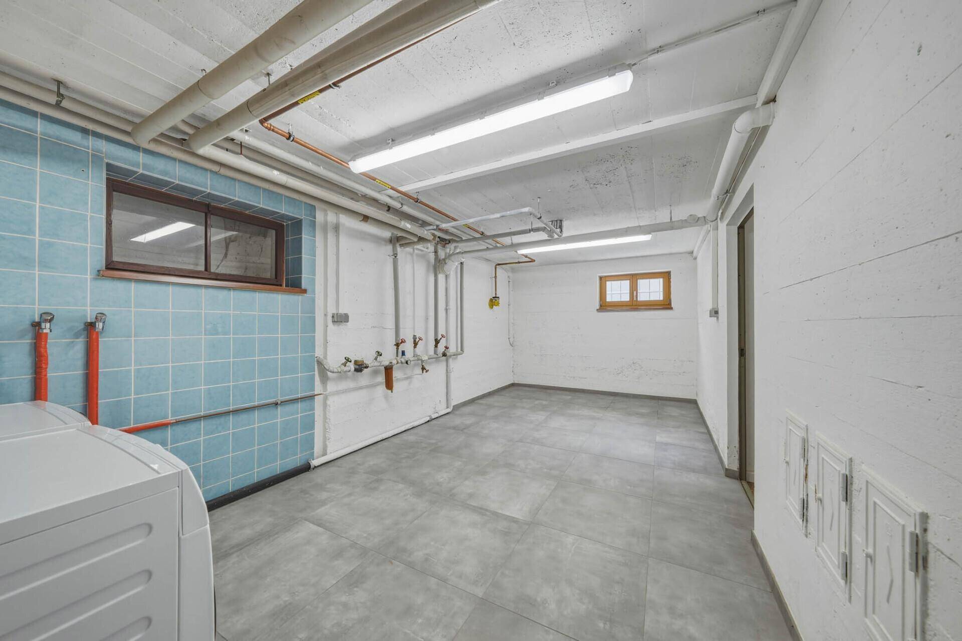 Hauswirtschaftsraum im Keller mit blauen Fliesen, sichtbaren Rohren und Waschmaschine.